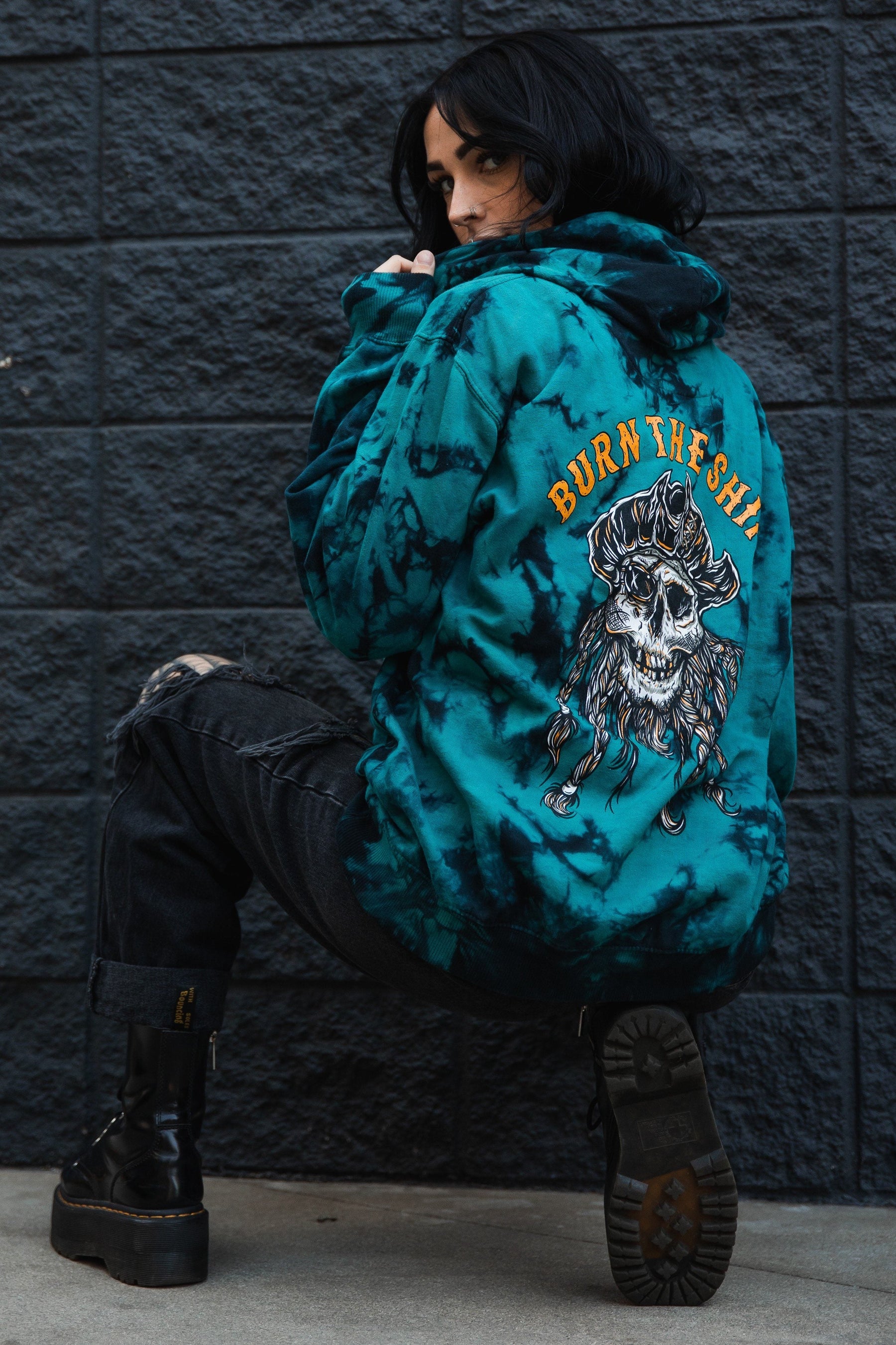 Someone in a Relentless Betrayal Burn The Ships tie dye hoodie, featuring a skull and crossbones design, crouches by a dark brick wall. They wear black ripped jeans and boots, with shoulder-length dark hair.