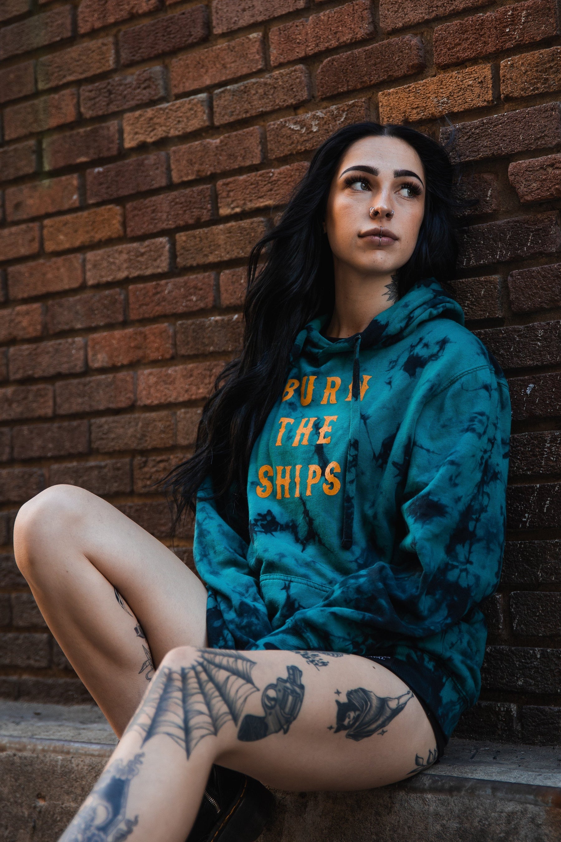 A woman with long dark hair sits against a brick wall wearing a Relentless Betrayal Burn The Ships Tie Dye Hoodie in blue, featuring BURN THE SHIPS in orange. The cozy cotton polyester blend complements her thoughtful expression, leg tattoos, and piercings.