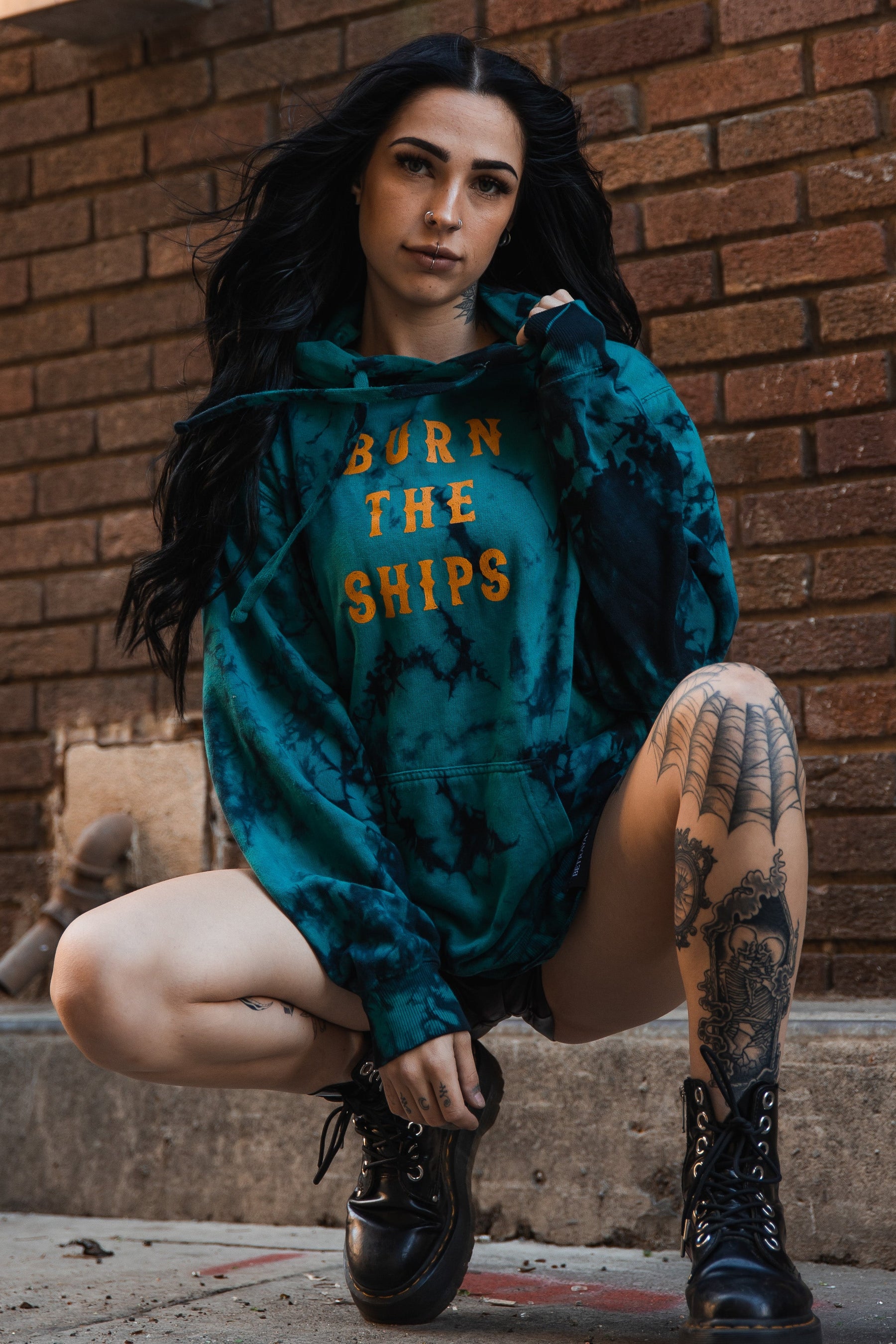 In front of a brick wall, a person with long dark hair squats in a blue tie-dye Burn The Ships hoodie by Relentless Betrayal, made from a cotton-polyester blend. Orange text stands out on the hoodie, while black boots and a tattoo on their left leg add to their urban vibe.