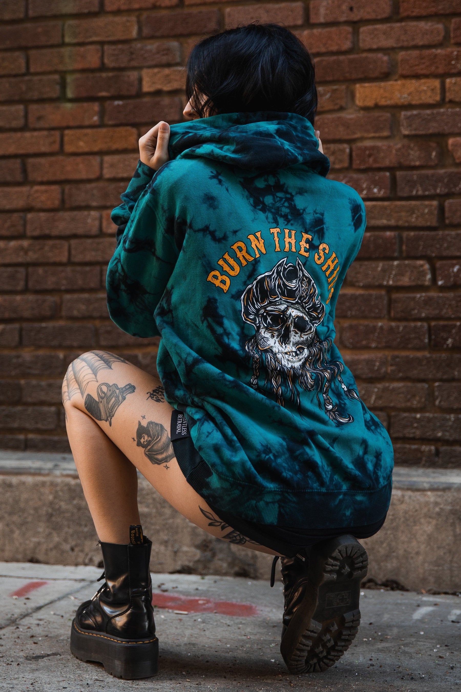 A person crouches on the sidewalk with their back to the camera, wearing a Relentless Betrayal Burn The Ships Tie Dye Hoodie made from a cotton-polyester blend, featuring a skull design. This unisex hoodie complements their tattooed legs and black boots against a brick wall backdrop.