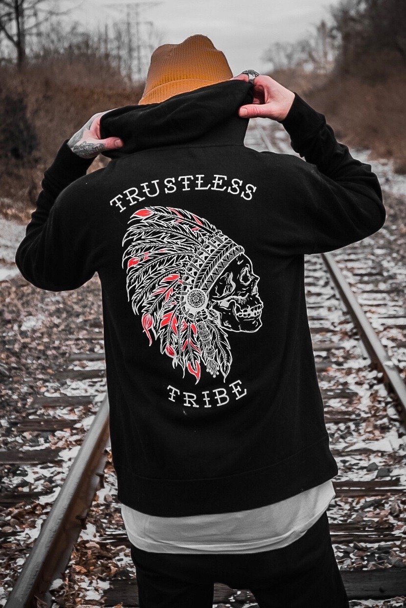A person wearing the Relentless Betrayal Trustless Chief Hoodie RED, featuring a unisex design with a striking skull in a feathered headdress and “Trustless Tribe” emblazoned on the back, stands on railroad tracks in a wintry, leaf-strewn setting. Crafted from a cozy cotton-polyester blend, this hoodie delivers both comfort and style even in the coldest weather.