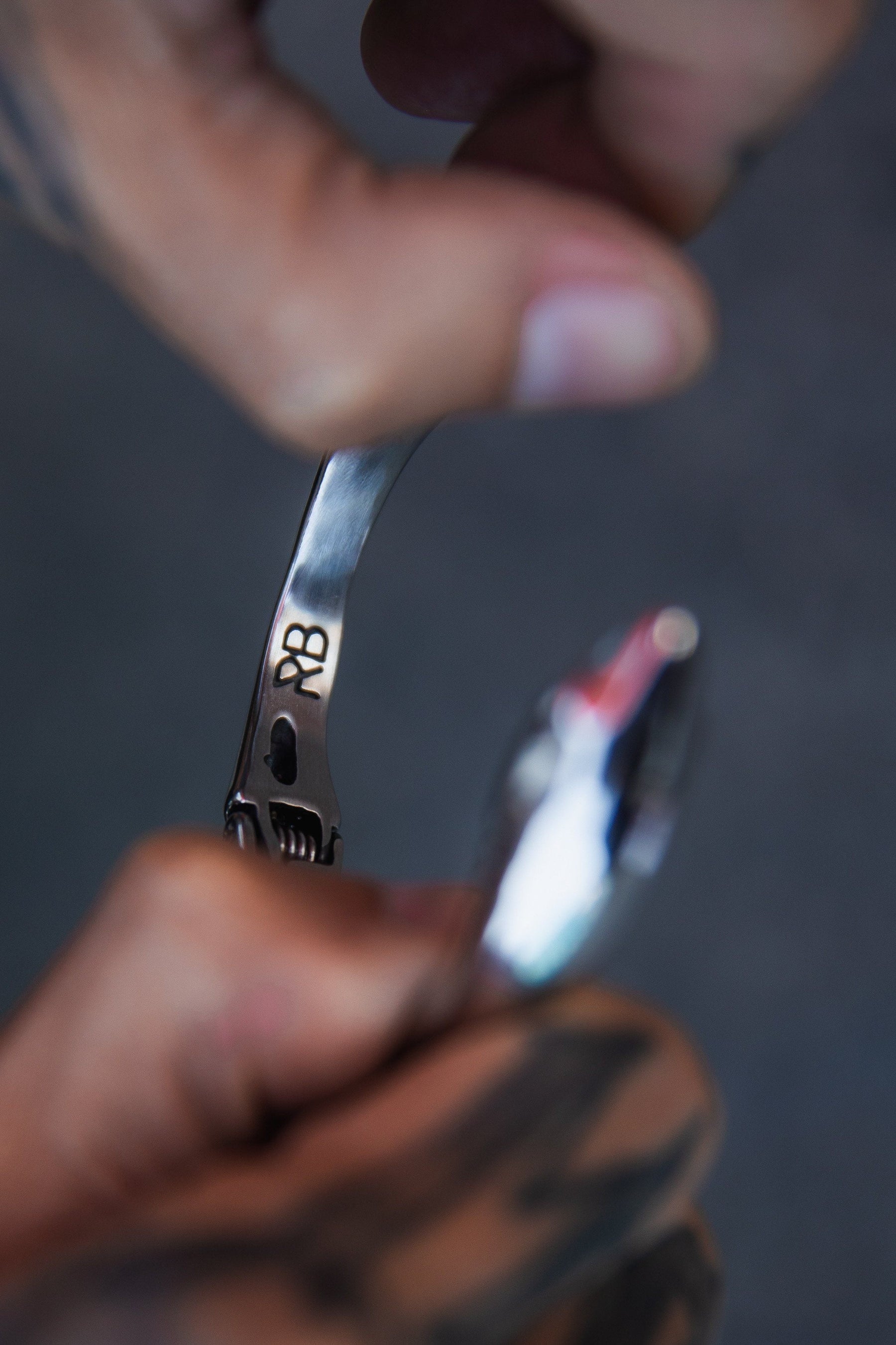 A tattooed person bends a stainless steel spoon by hand. The handle has RB engraved, representing Relentless Betrayal. The blurred background emphasizes the spoon and hands, echoing the creation of a Shark Bait Bracelet.