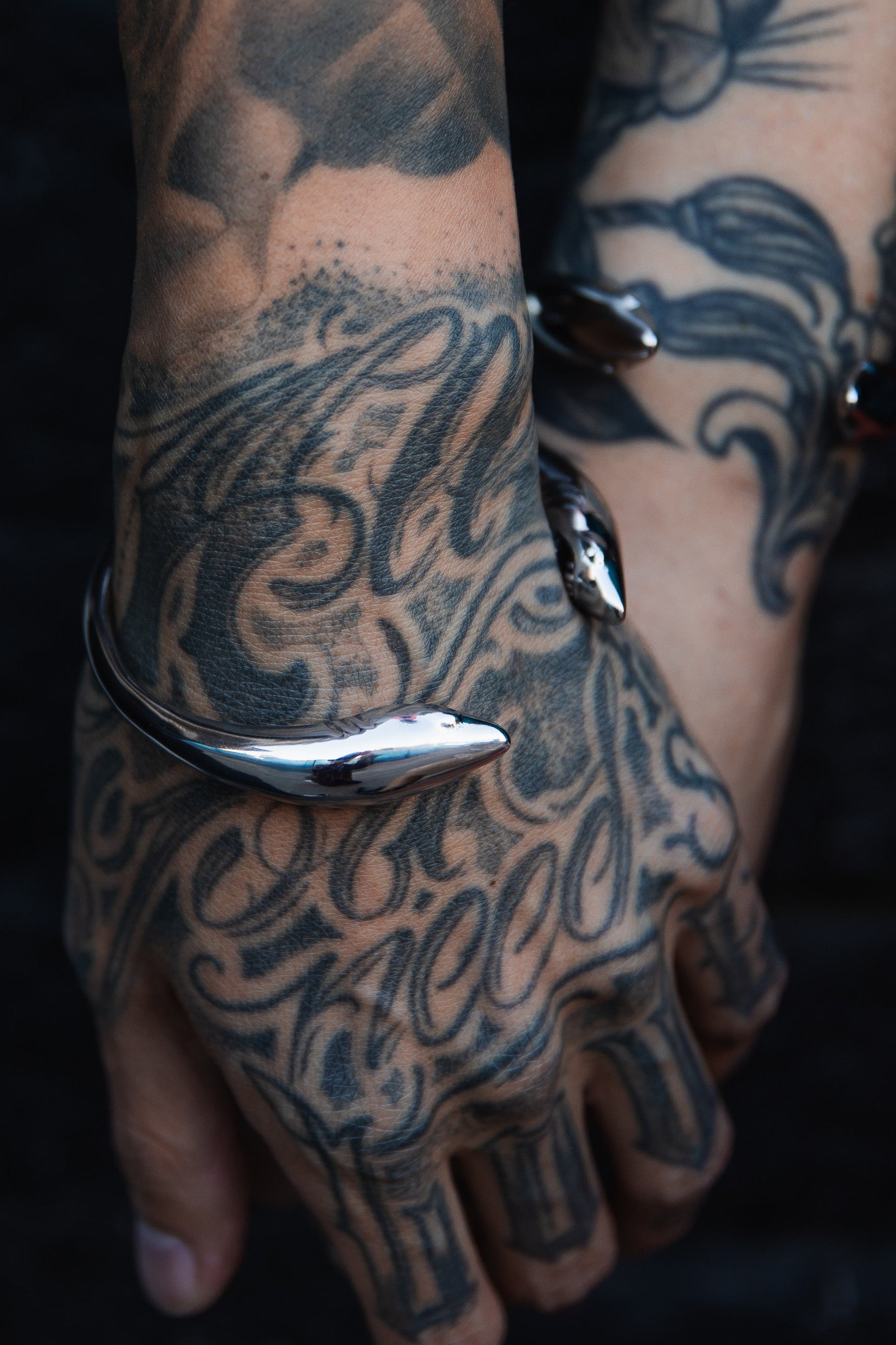 Tattooed hands with intricate scripts and designs are set against a dark background. On one wrist, a shiny stainless steel Shark Bait Bracelet by Relentless Betrayal shines, providing a striking contrast to the detailed tattoos.