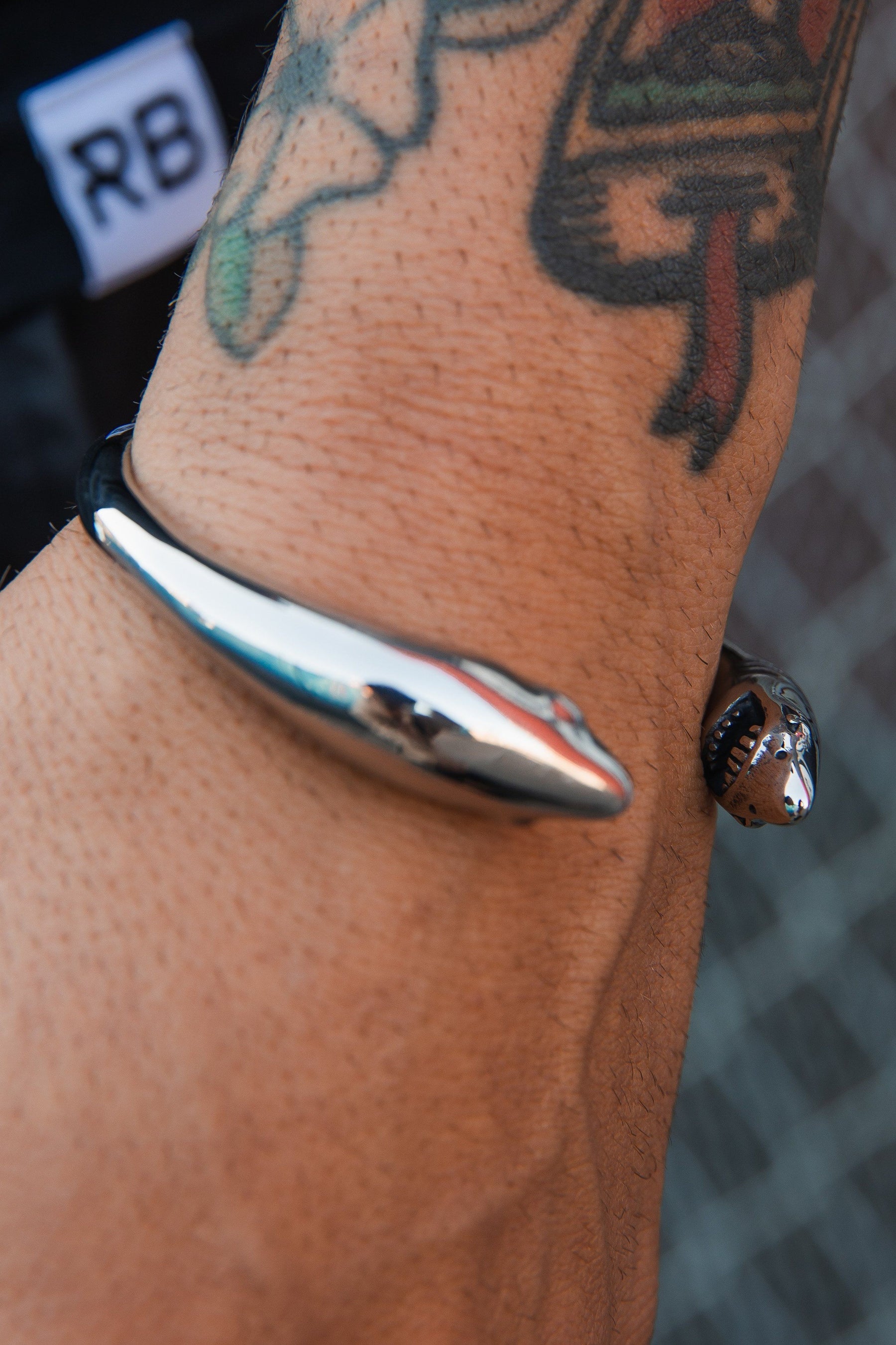 The Relentless Betrayal Shark Bait Bracelet is a stainless steel piece designed with opposing snake heads. It elegantly wraps around a tattooed wrist, its silver shine contrasting with vibrant ink against a blurred chain-link fence, and promises durability with its lifetime warranty.