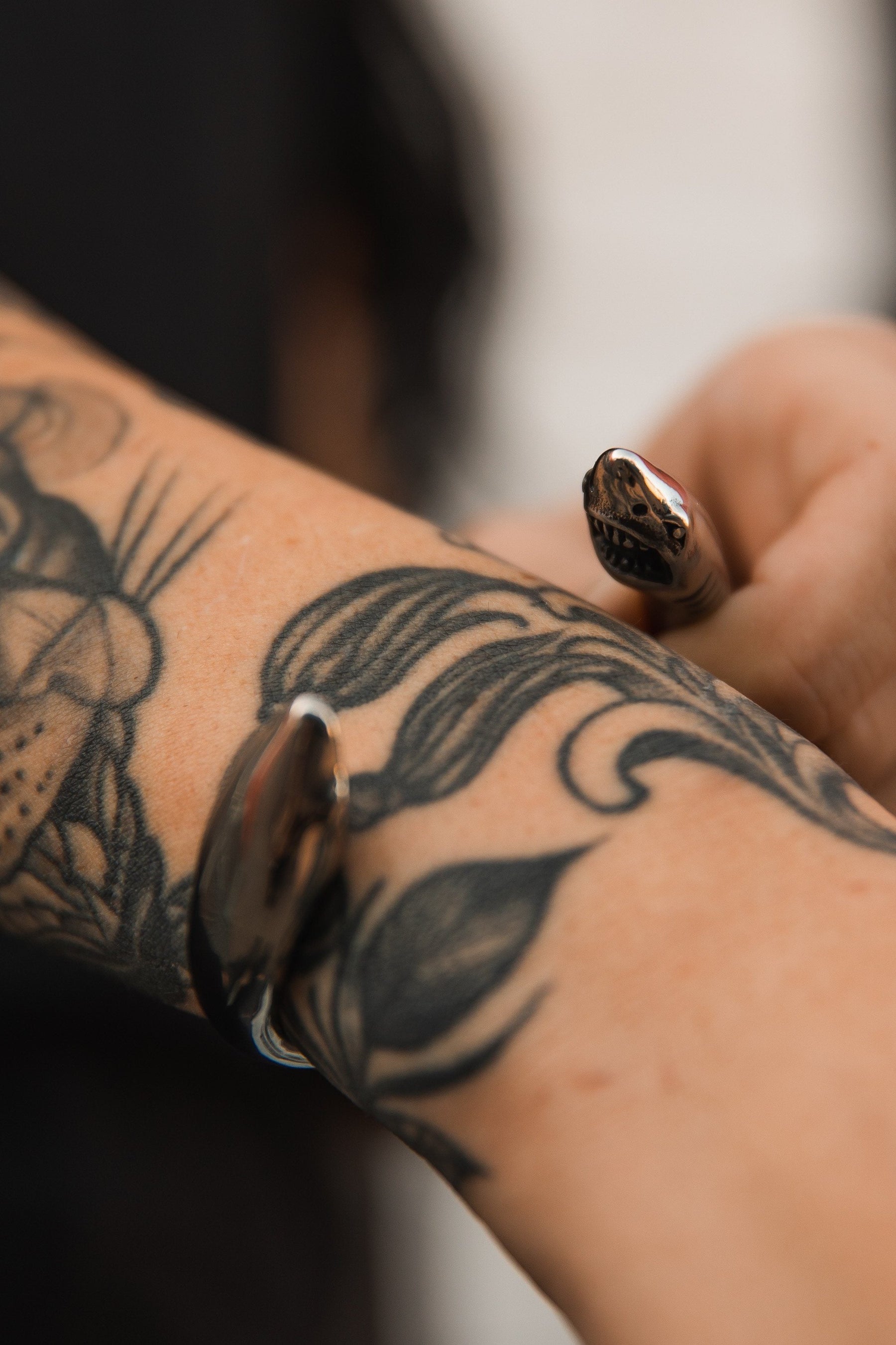 A detailed tattoo of leaves and flowers adorns the forearm, while a hand models Relentless Betrayals Shark Bait Bracelet near the wrist. The blurred background emphasizes the art and jewelry.
