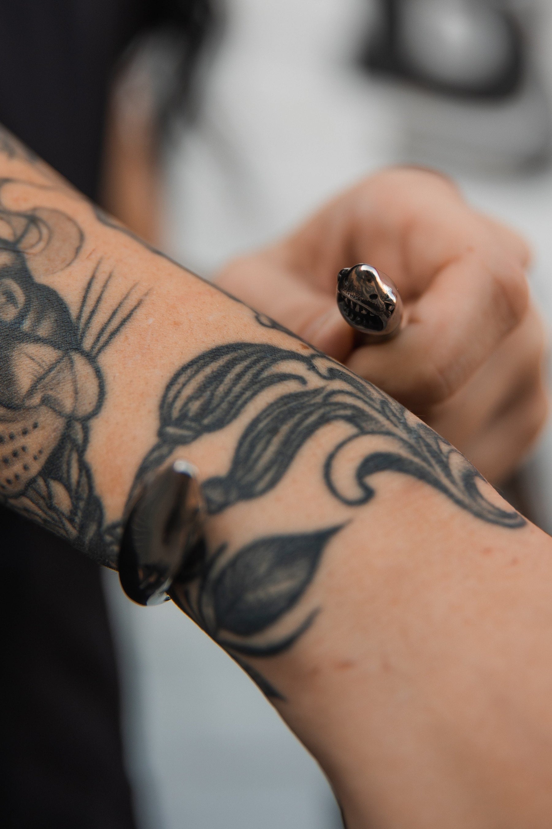 A close-up of a forearm shows a floral tattoo and the striking Relentless Betrayal Shark Bait Bracelet. Crafted from stainless steel with snake head shapes, this metallic accessory offers timeless appeal against a blurred backdrop.