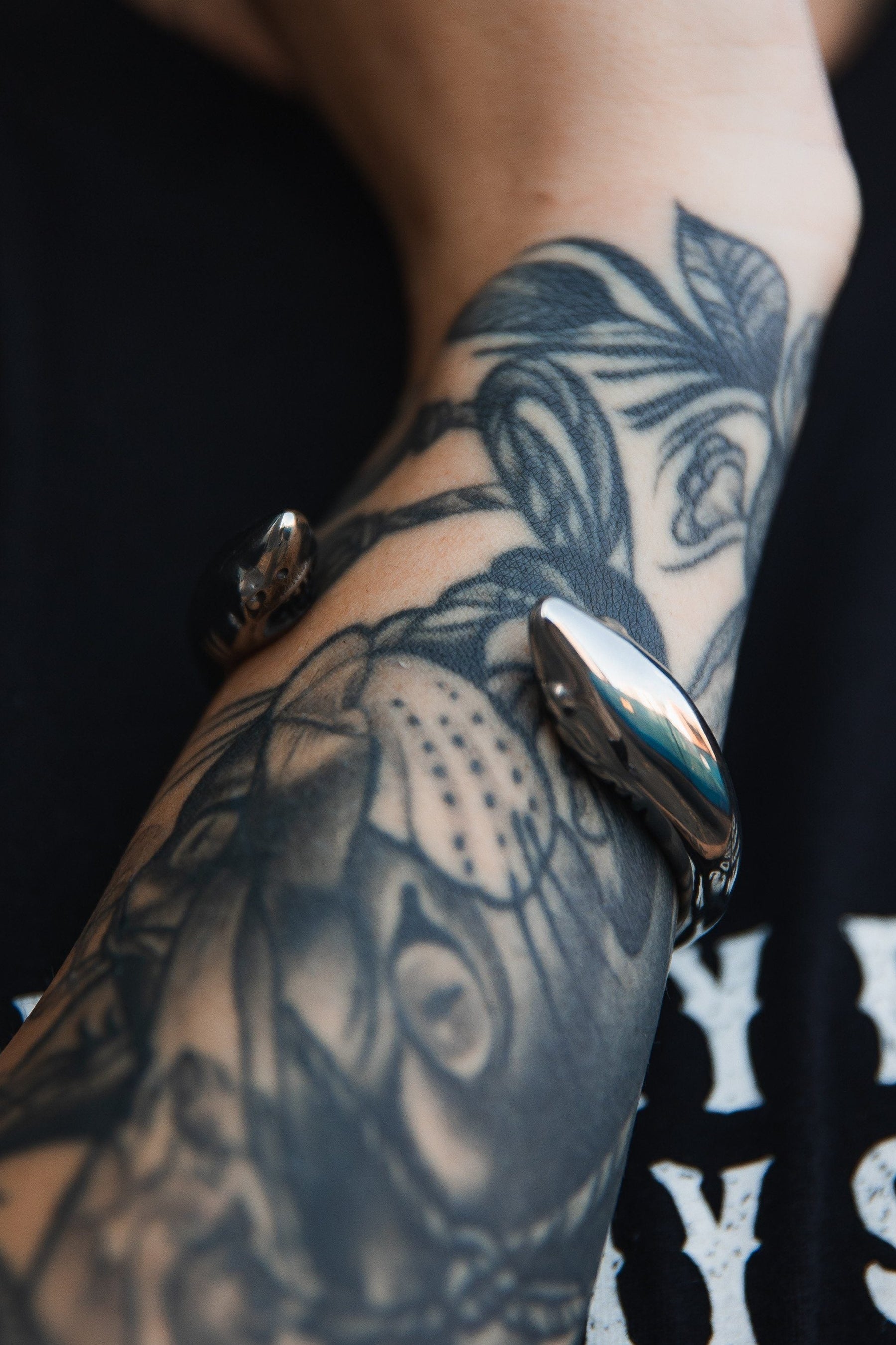 A close-up of a tattooed forearm is elegantly adorned with the Shark Bait Bracelet by Relentless Betrayal, its stainless steel design gleaming against black-and-white floral and animal tattoos. The backdrop is a black T-shirt with partial white text.