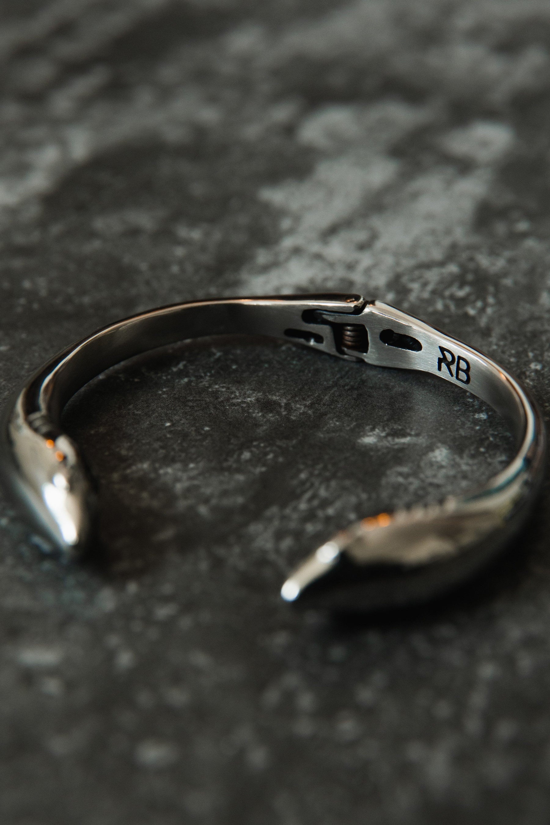 The Relentless Betrayal Shark Bait Bracelet, in elegant silver stainless steel with arrow-shaped ends and an RB engraving, rests stylishly on a textured dark surface. It includes a lifetime warranty for added assurance.