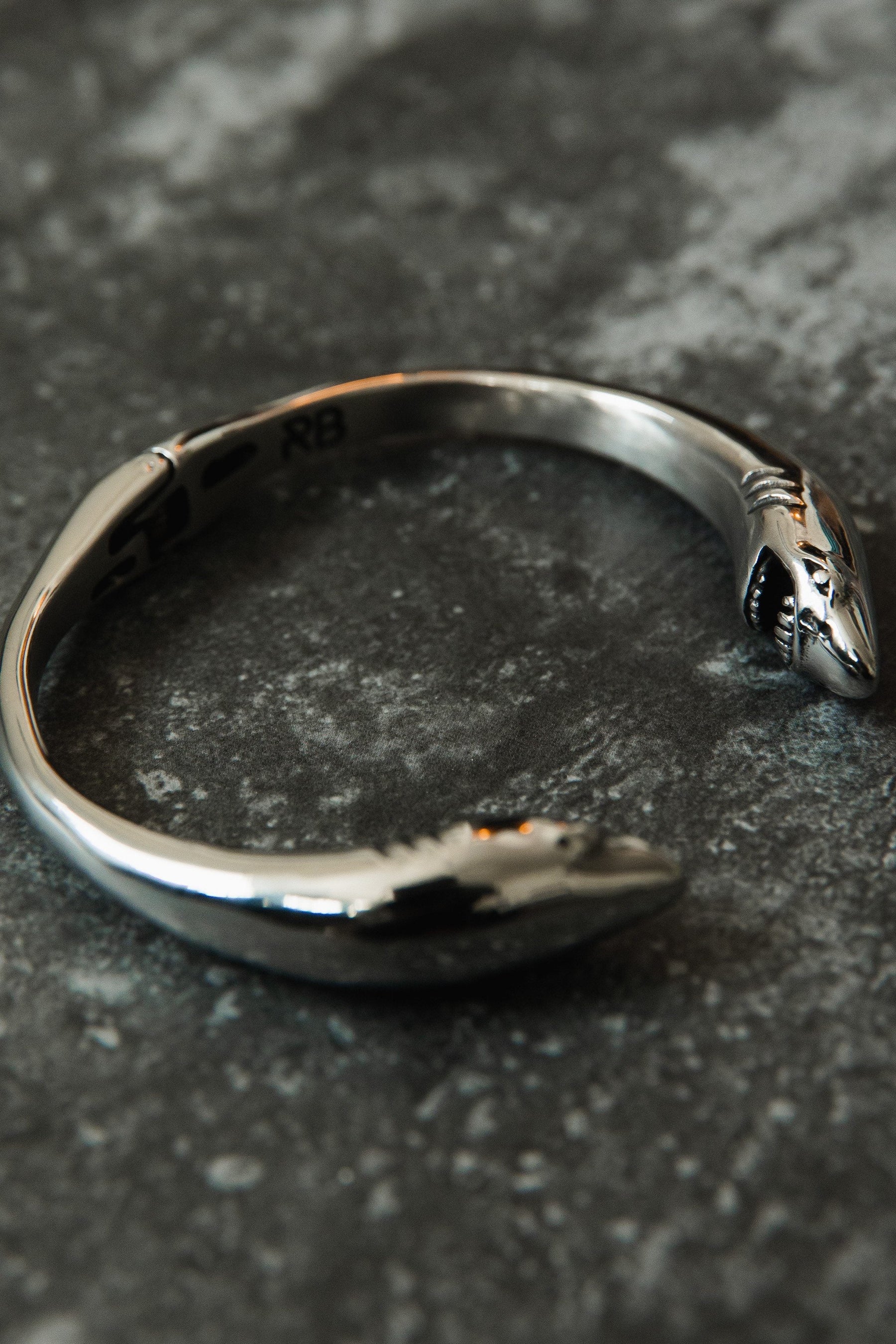 Introducing the Shark Bait Bracelet by Relentless Betrayal, made from stainless steel and featuring two distinctive shark heads. Its textured dark gray surface offers a soft shine, and it comes with a lifetime warranty for added peace of mind.