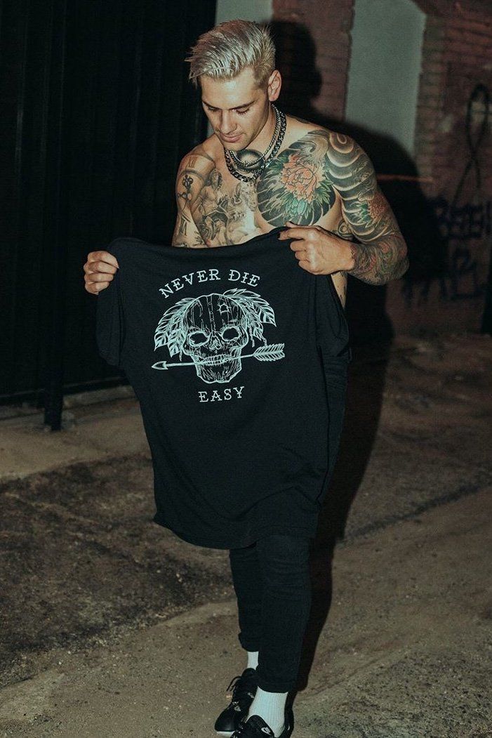 A tattooed man on a night street wears the Relentless Betrayal Never Die Easy Black Tee, featuring a skull and crossed arrows. Made from fine jersey cotton, this unisex tee pairs perfectly with his dark pants and shoes.