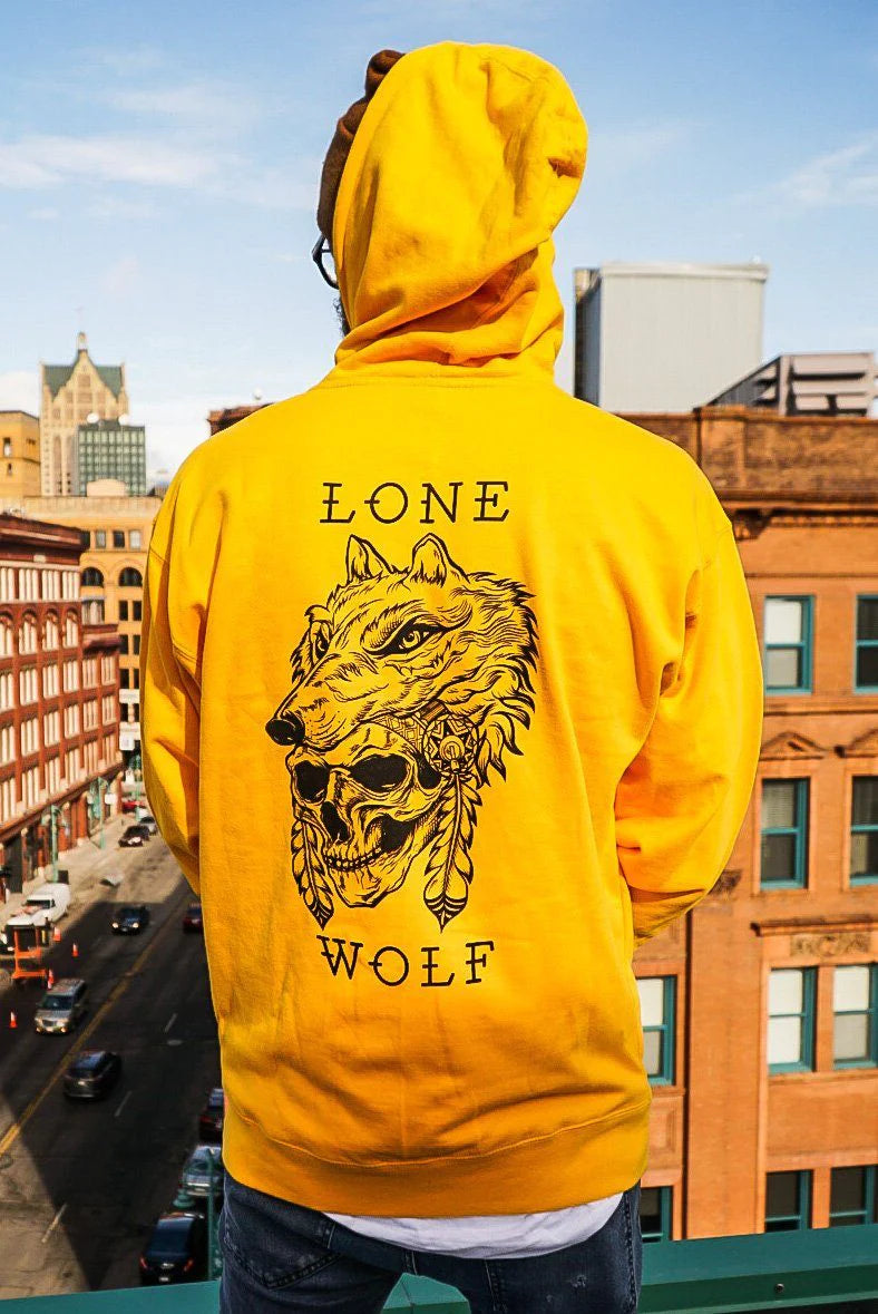 A person in a Relentless Betrayal Lone Wolf GOLD Hoodie stands on a rooftop overlooking city streets. The unisex hoodie showcases a graphic of a wolf with a headdress and the text Lone Wolf, with buildings and clear skies in the background.