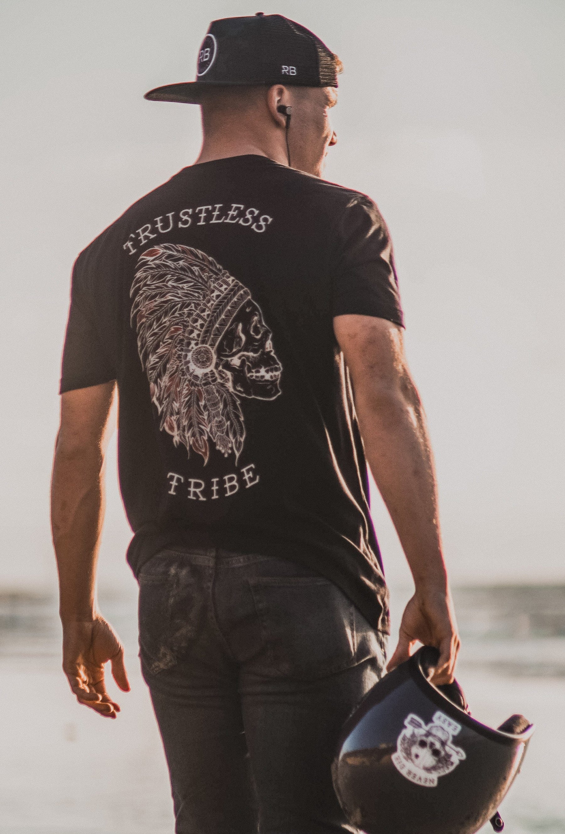 A man in a black cap and the Relentless Betrayal Trustless Chief Tee - Red with a skull and headdress design stands on the beach. Channeling his inner rebel leader, he holds a helmet with the same logo, gazing towards the ocean under the clear sky.