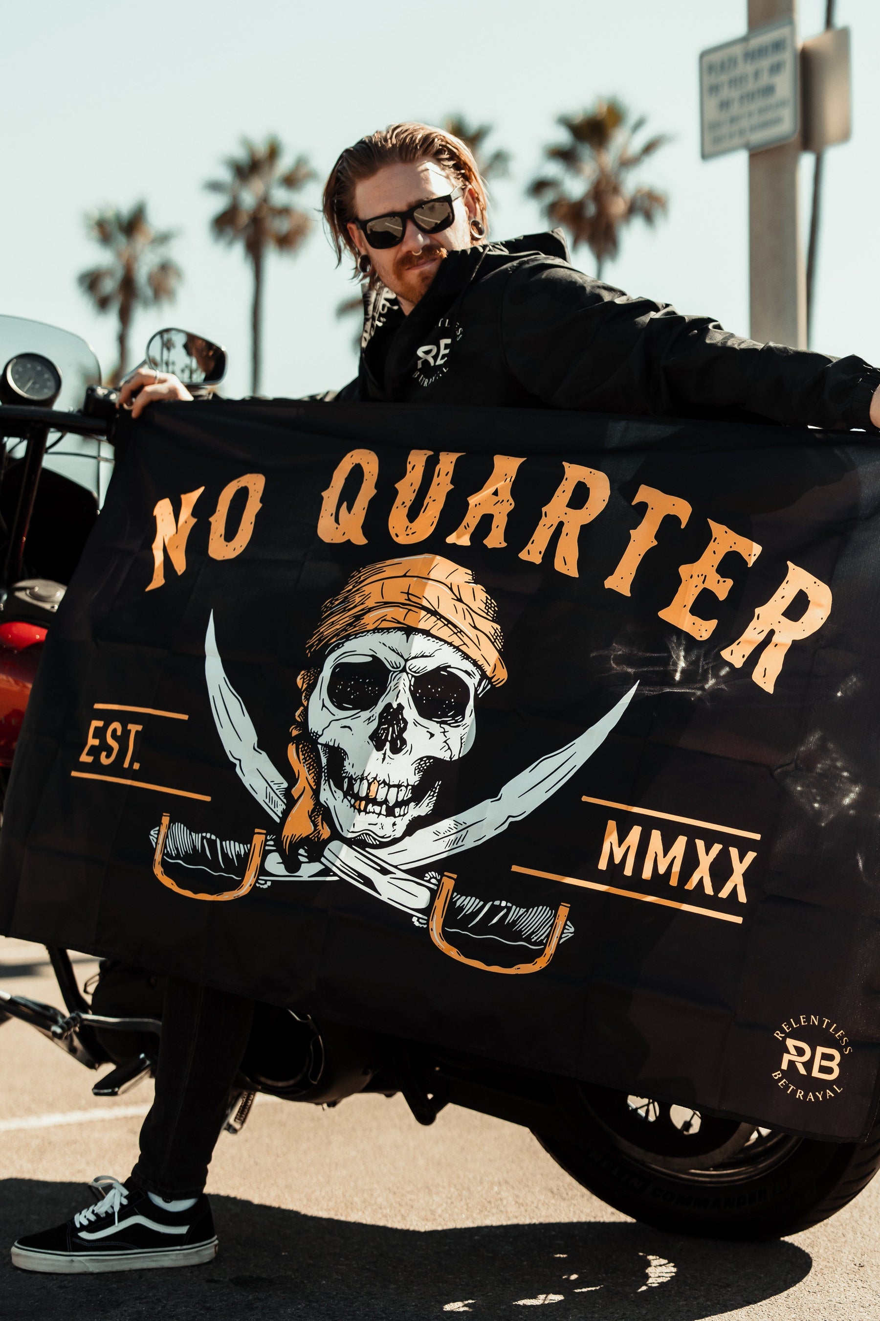A person in sunglasses and a leather jacket stands next to a motorcycle, holding the Relentless Betrayals No Quarter Flag, made of durable polyester. The flag depicts a skull with a bandana, crossed swords, and No Quarter Est. MMXX text as palm trees sway gently in the background.