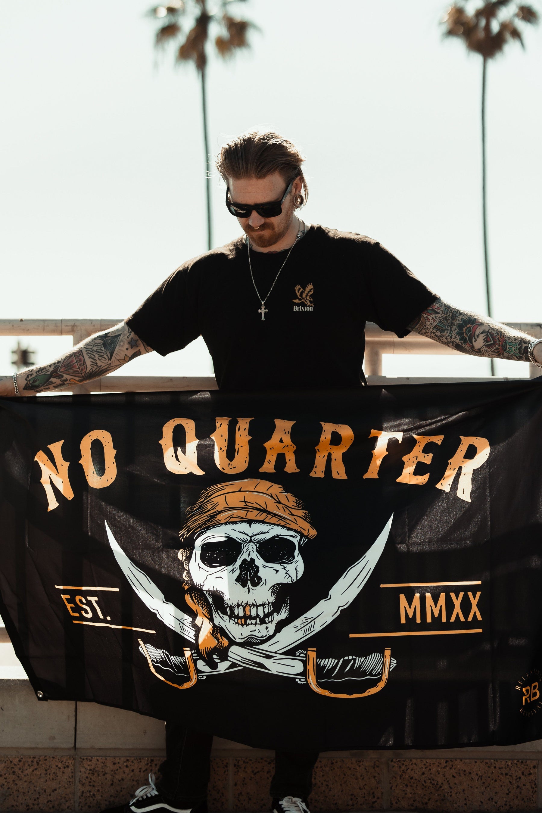 A tattooed person wearing sunglasses holds the Relentless Betrayals No Quarter Flag, a full-size pirate-themed banner made of durable polyester. It features a skull with a bandana above crossed swords, waving against palm trees and a railing.