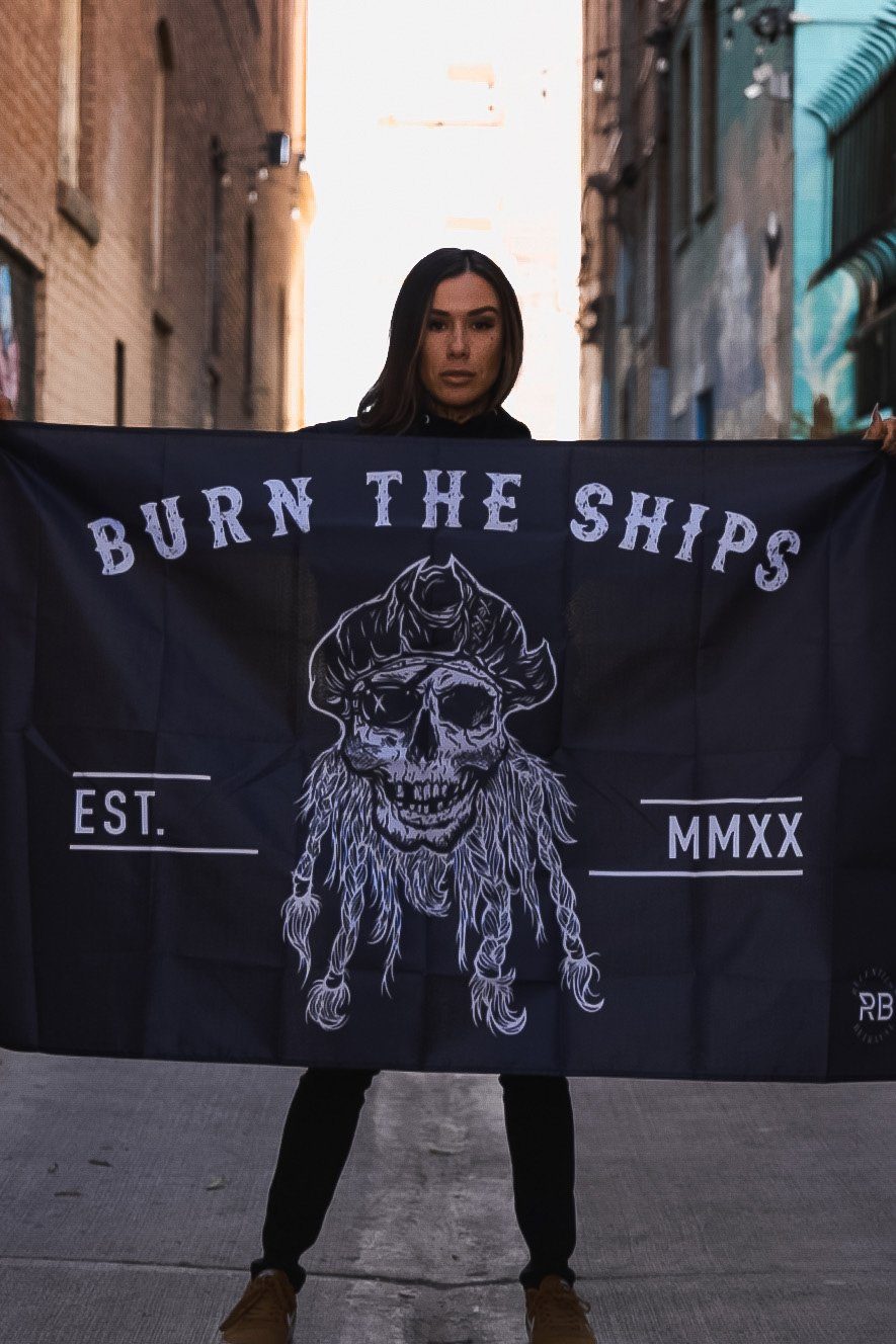 In an alley, a person waves the Relentless Betrayals Burn The Ships Flag, made of durable polyester and emblazoned with a skull and crossbones. BURN THE SHIPS and EST. MMXX stand out as it flutters among brick and metal buildings, held by sturdy metal grommets.