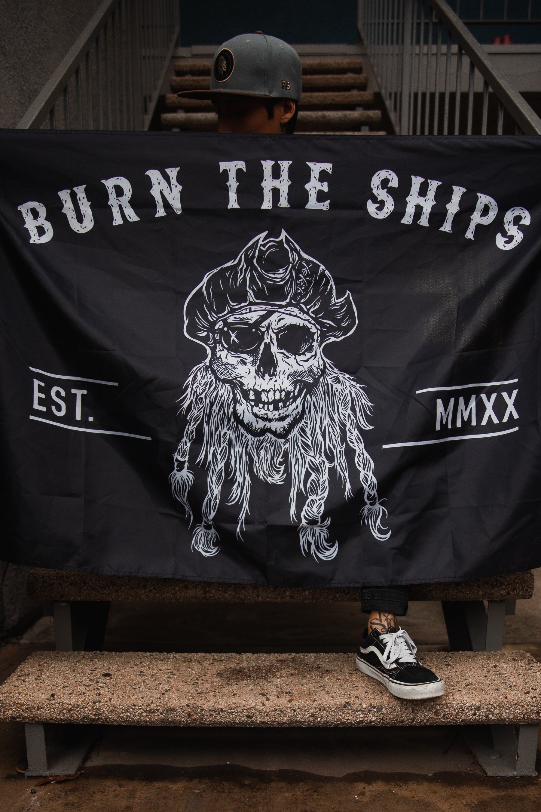 A person sits on concrete steps holding the Burn The Ships Flag by Relentless Betrayal, crafted from durable polyester. This black flag features a skull with a tricorne hat and beard, BURN THE SHIPS above, EST. MMXX on the sides, and metal grommets for durability.