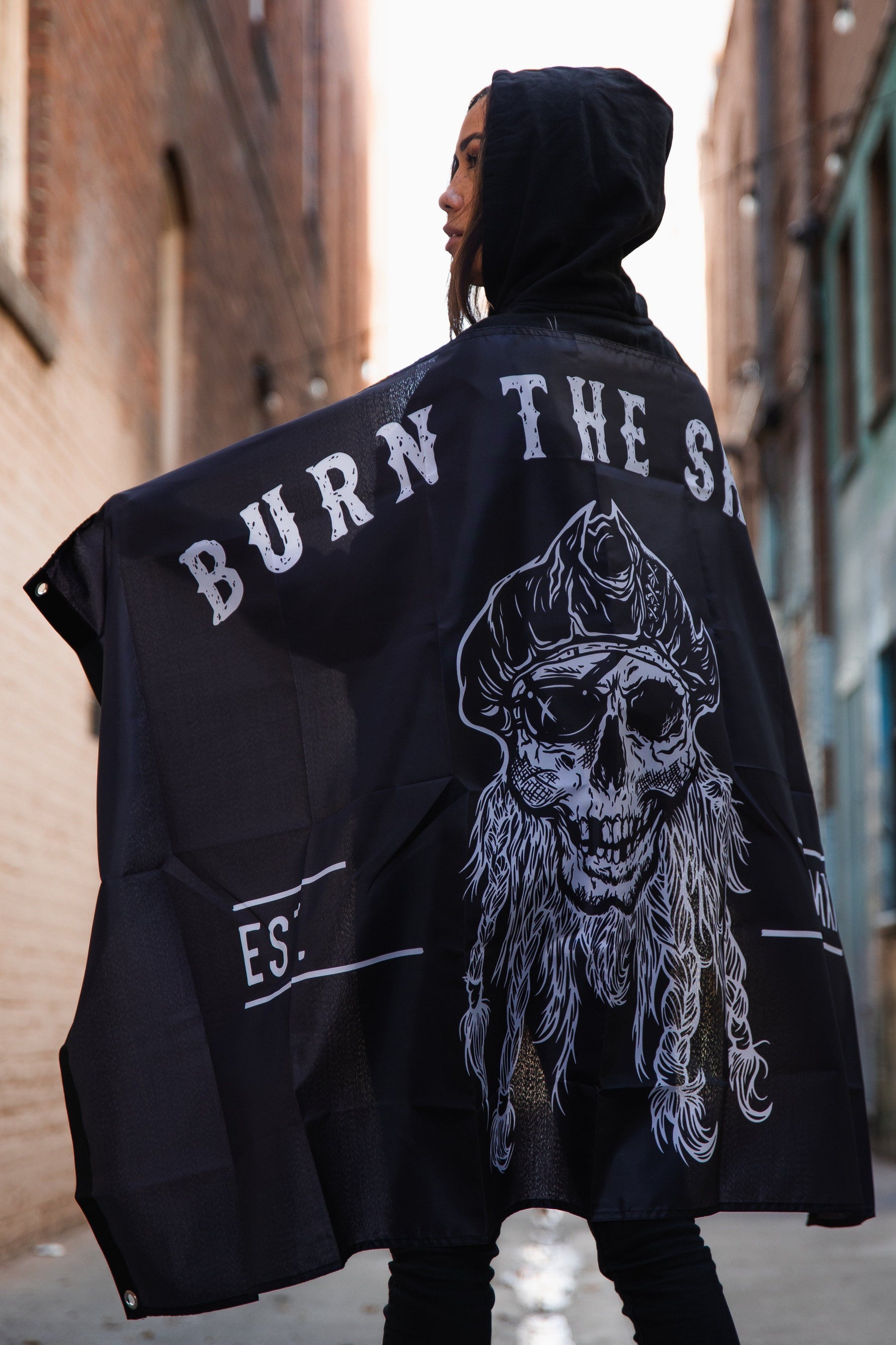 In a shadowy alleyway, a hooded figure raises the Burn The Ships Flag by Relentless Betrayal, made of durable polyester with BURN THE SHIP and a skull design. Metal grommets enhance its strength as dim light filters down the narrow, brick-lined path.