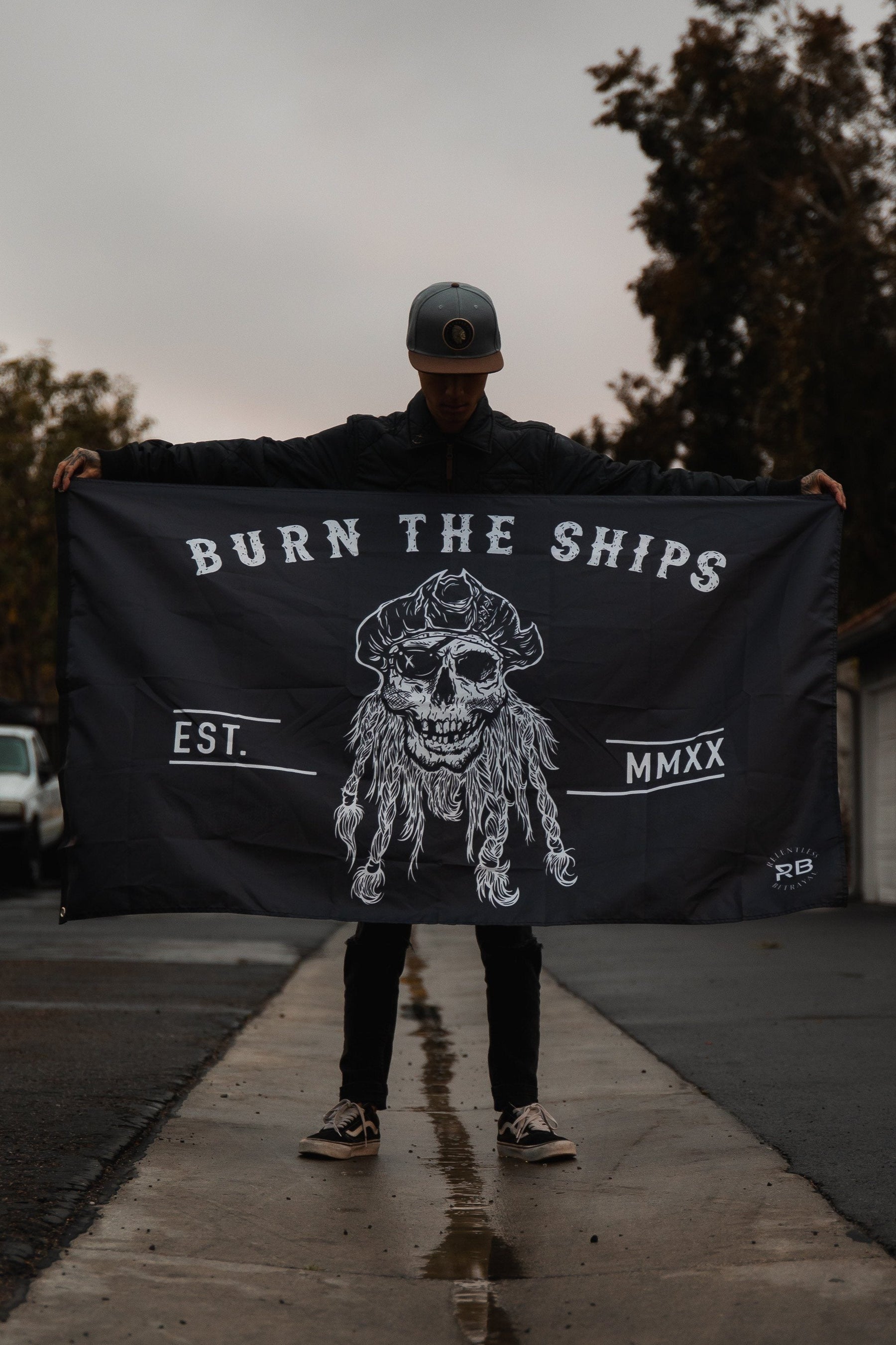 On the wet pavement, a person holds the Burn The Ships Flag by Relentless Betrayal, displaying a skull with BURN THE SHIPS, EST. MMXX, and RB. Metal grommets glint subtly under the hazy sky framed by distant trees. The person wears a cap, standing defiantly.