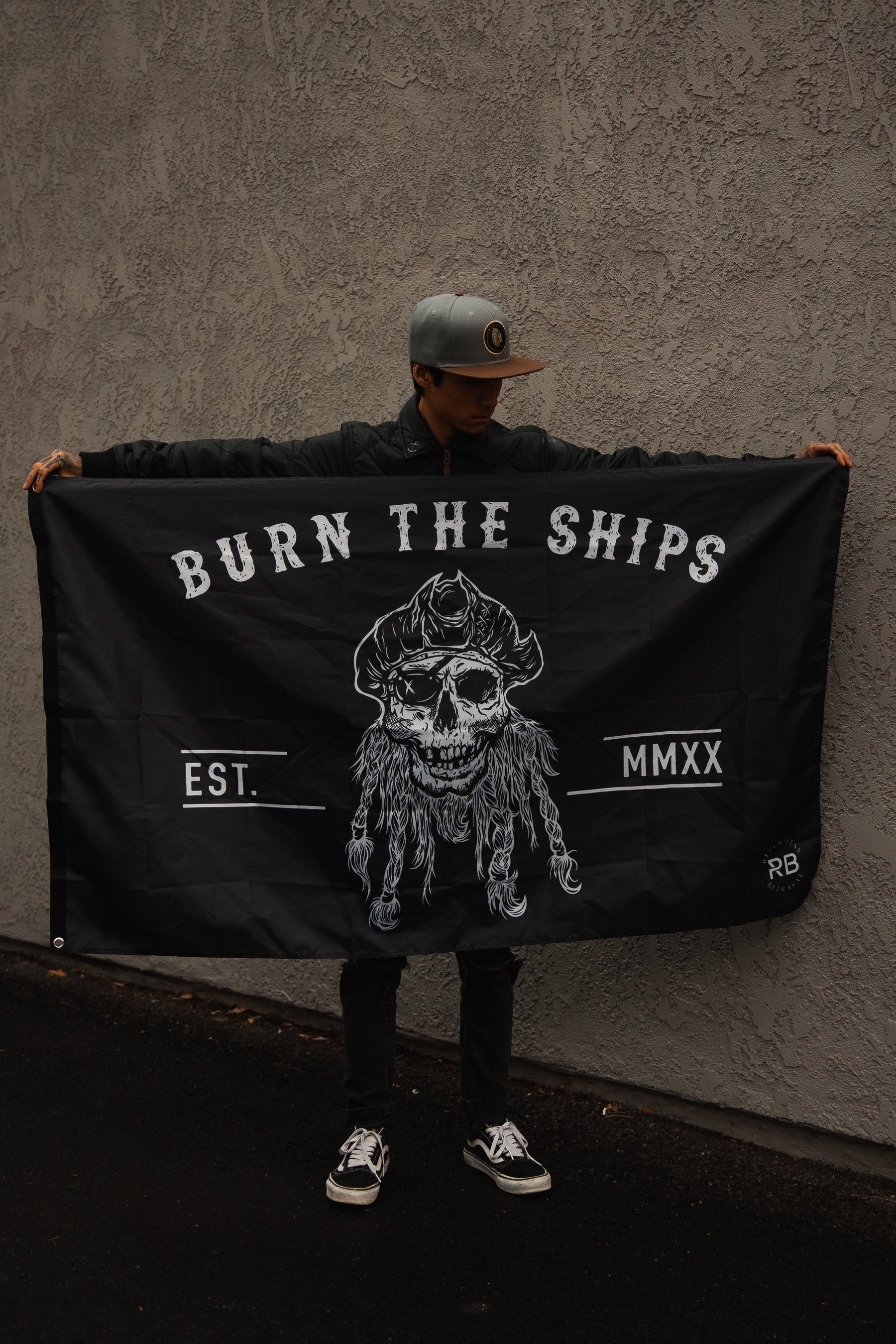 A person confidently stands with a Relentless Betrayals Burn The Ships flag featuring a skull, EST. MMXX, in durable polyester with metal grommets, against a textured gray wall, while wearing a cap, jacket, and sneakers.