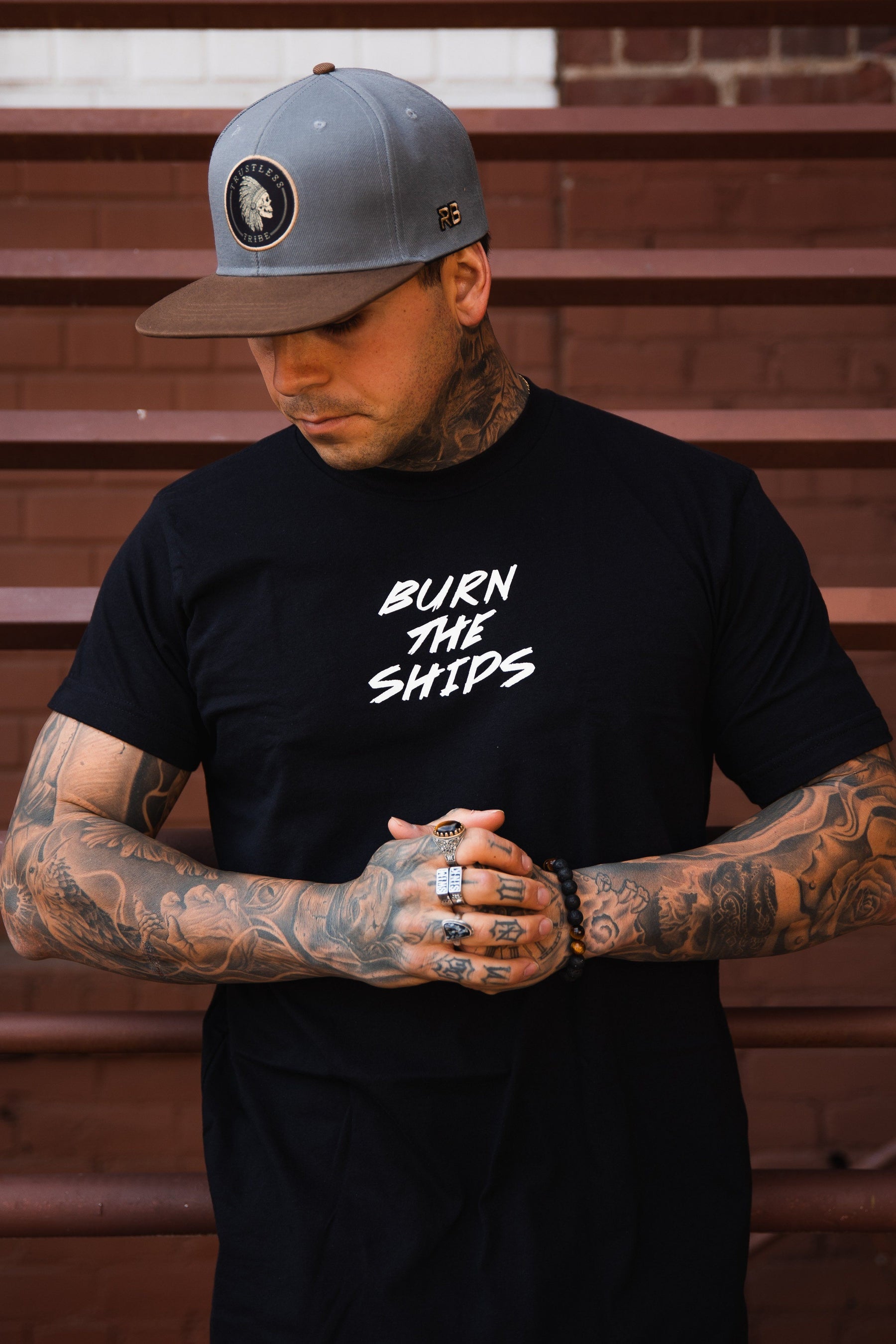 A tattooed man wearing a Relentless Betrayal Burn The Ships Traditional Tee and a gray cap stands with clasped hands. He wears multiple rings and a bracelet, set against a brown brick wall and stairs.
