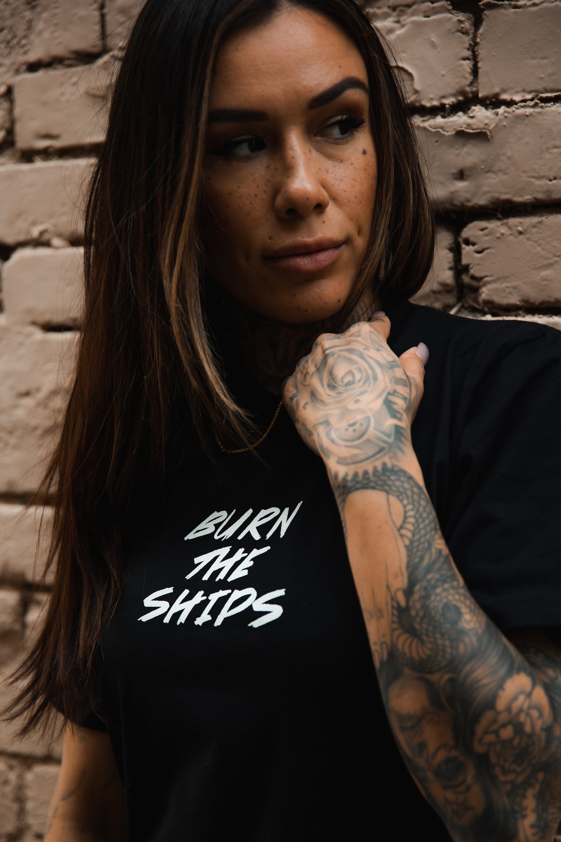 A long-haired, tattooed individual stands by a brick wall, wearing the Burn The Ships Traditional Tee from Relentless Betrayal. The unisex black tee, made of fine jersey cotton, features the Burn the Ships print. They gaze to their left.