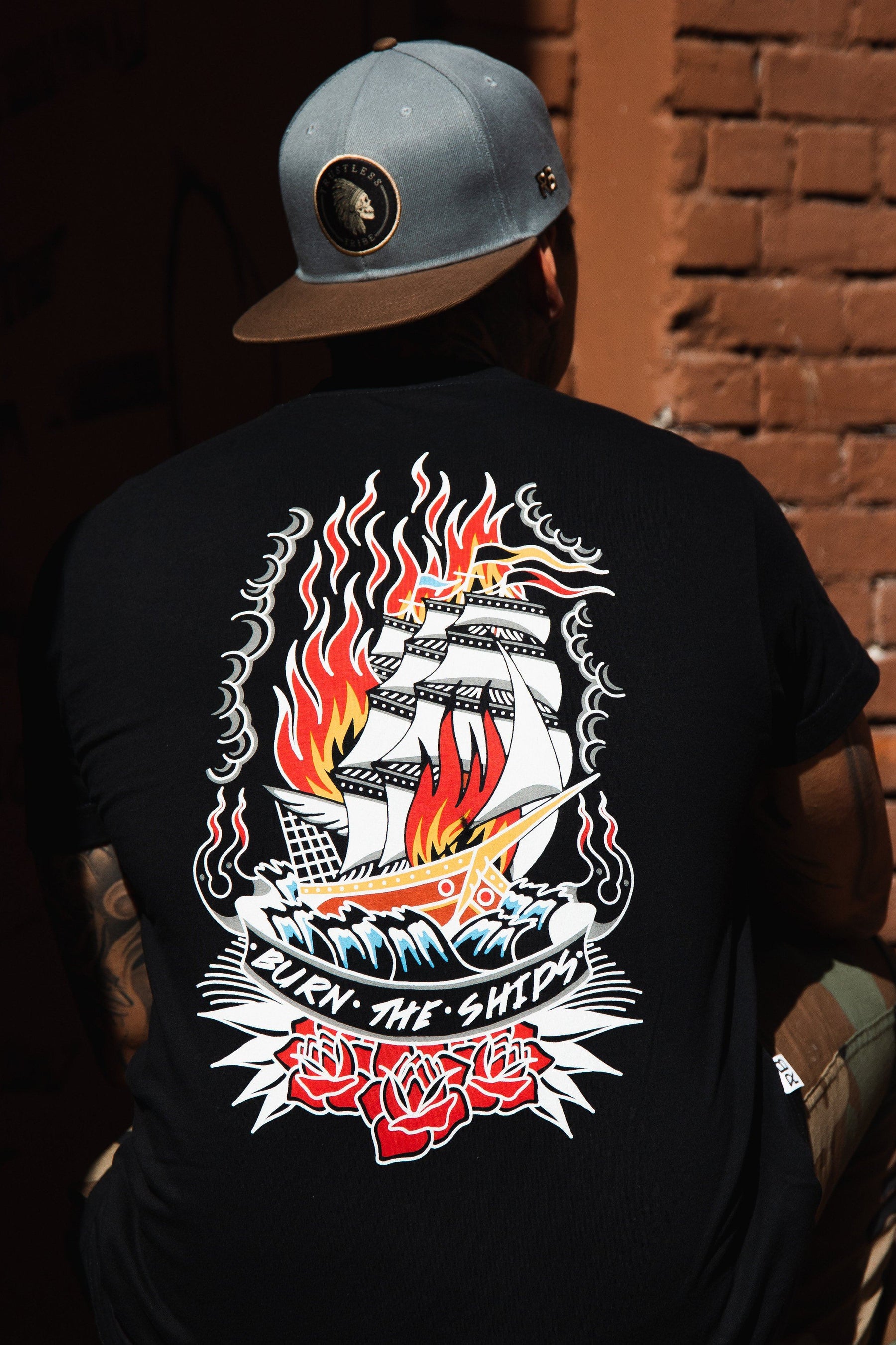 Sporting a Burn The Ships Traditional Tee by Relentless Betrayal, featuring vibrant graphics of a ship with flames and roses and the phrase Burn the Ships, a person wears this unisex fine jersey cotton T-shirt, double-stitched for durability, along with a gray cap in front of a brick wall.