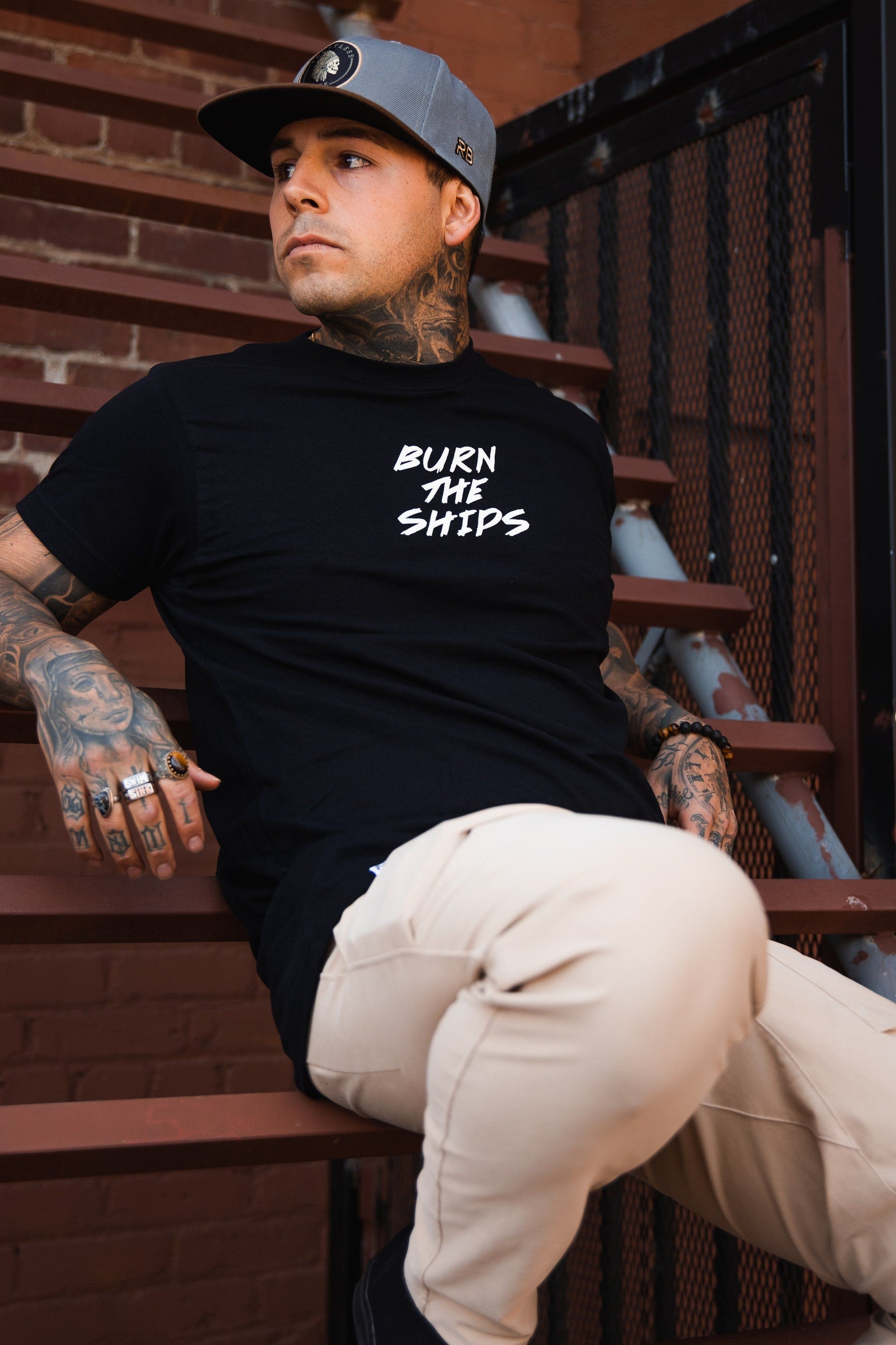 A tattooed man, wearing a gray cap and the Burn The Ships Traditional Tee by Relentless Betrayal in black fine jersey cotton, sits on metal stairs. Dressed in beige pants, he thoughtfully gazes to the side against a brick wall backdrop.