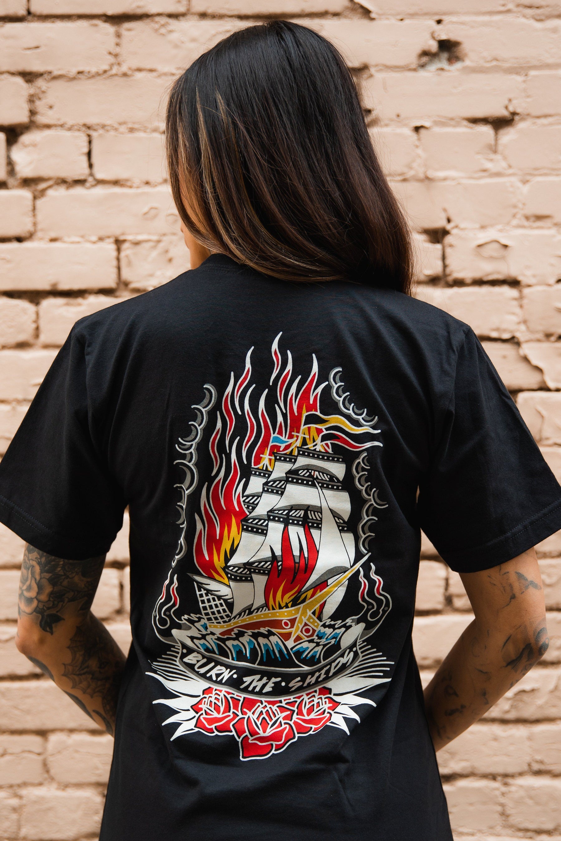 A person with long hair and tattooed arms faces away, wearing the Relentless Betrayal Burn The Ships Traditional Tee, featuring a flaming ship with red roses. Made from fine jersey cotton with a double-stitched design, it stands out against a light brick wall.