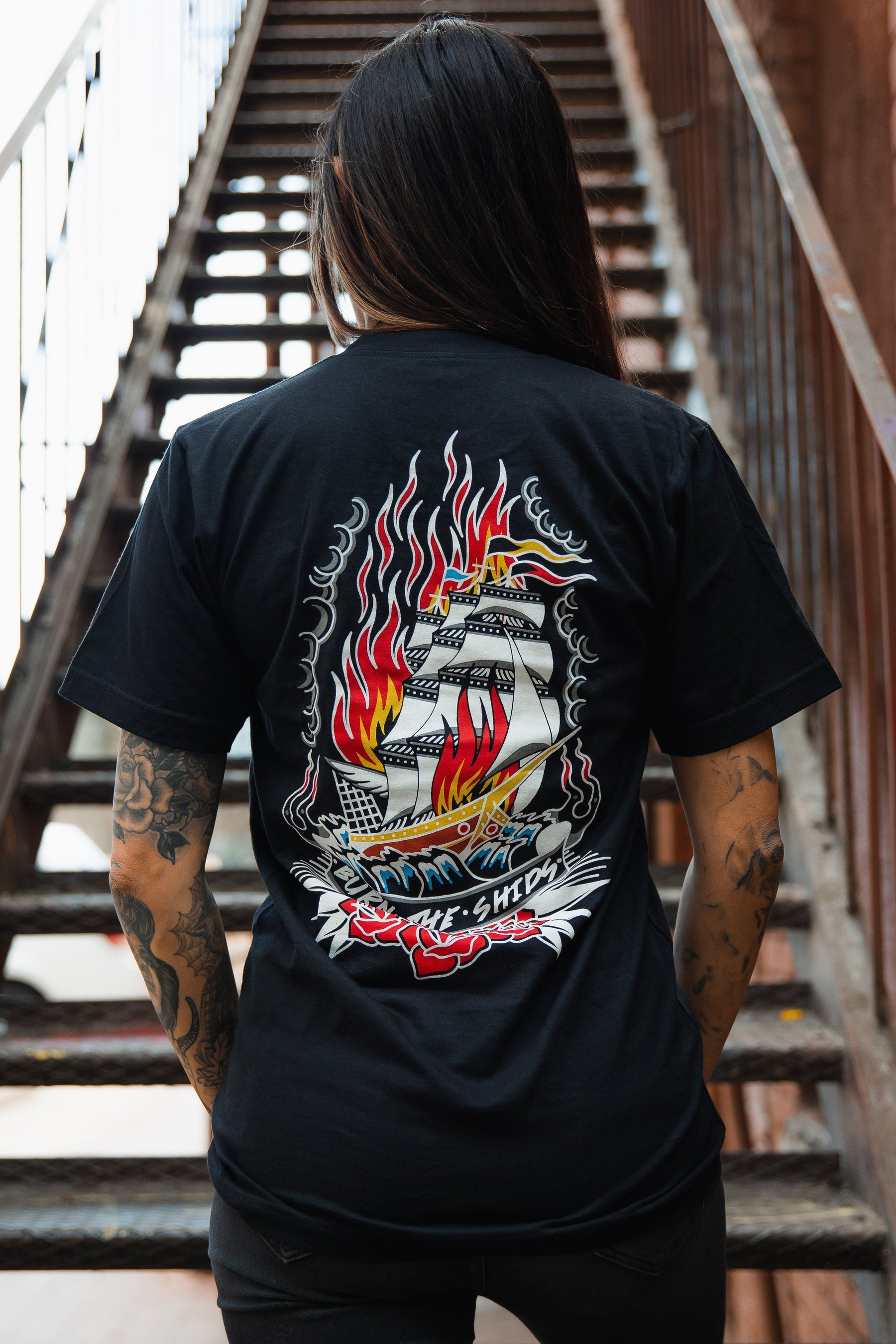 Standing on a staircase with long hair and tattoos, the individual sports a Relentless Betrayal Burn The Ships Traditional Tee, a black unisex fine jersey cotton shirt featuring a vibrant ship-in-flames graphic and double-stitched design.