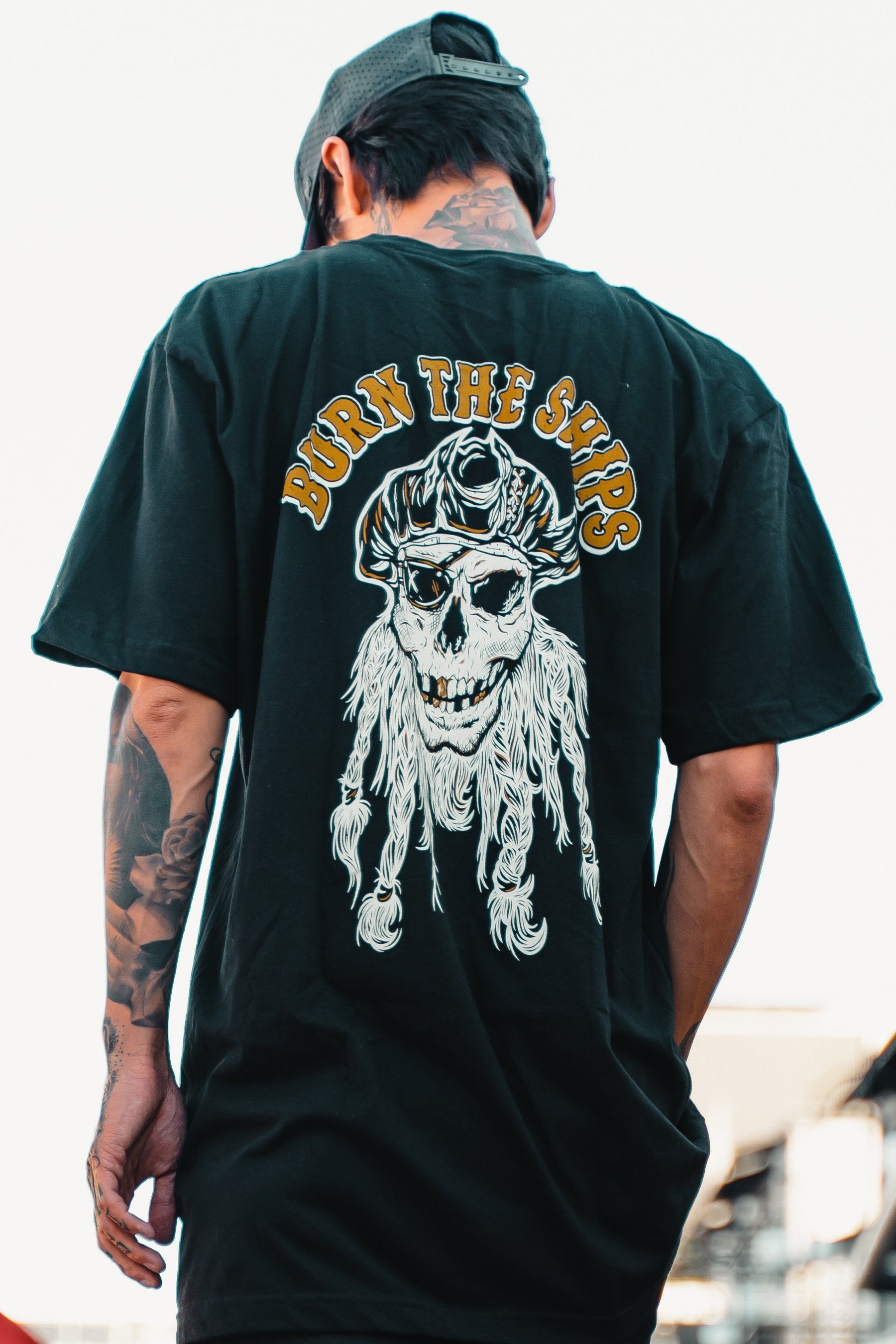 A person in a Burn The Ships Gold & White Tee by Relentless Betrayal, featuring a pirate skull design and BURN THE SHIPS in bold yellow, showcases their tattoos while wearing a backward cap against a bright, slightly blurred background for an edgy vibe.