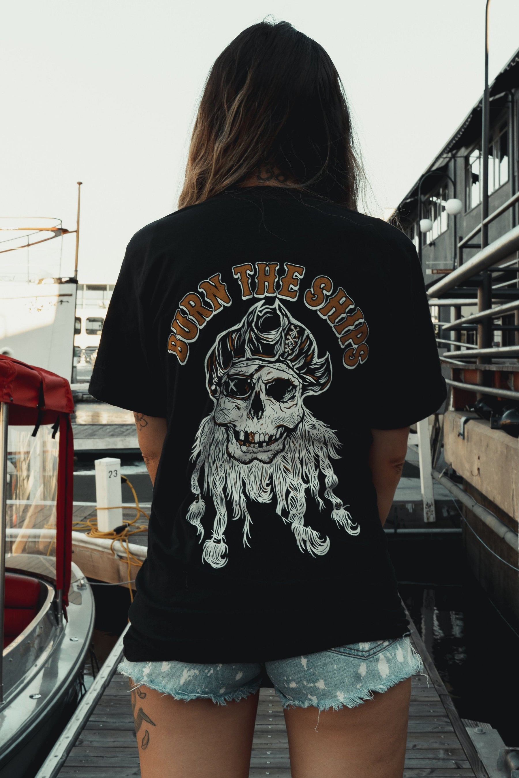 A person stands on a dock wearing Relentless Betrayals Burn The Ships Gold & White Tee, a black cotton shirt featuring a pirate-themed skull design with the slogan. Frayed denim shorts complete the look, set against a backdrop of boats and buildings.