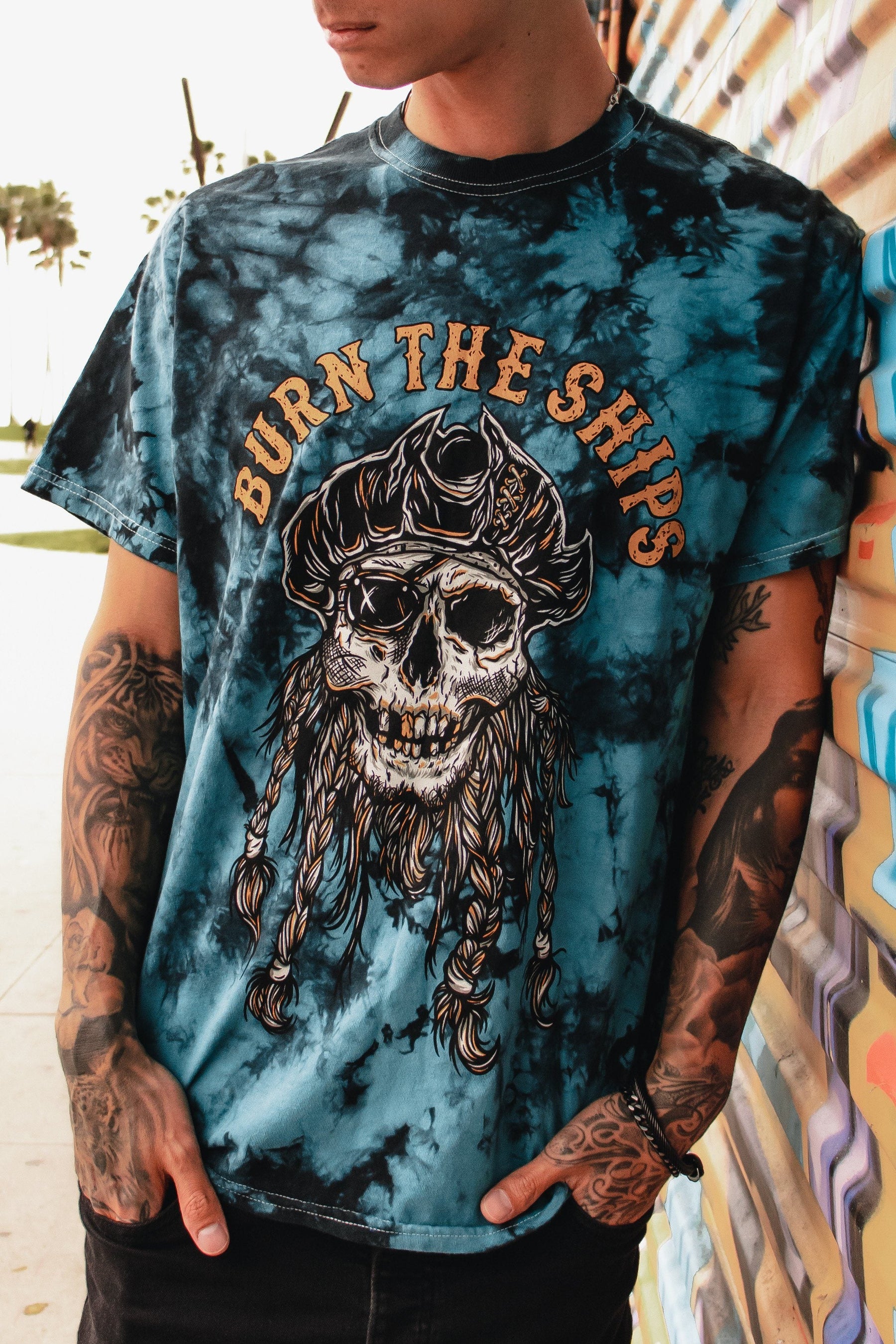 A person in a Relentless Betrayal Burn The Ships Tie Dye Tee, showcasing a pirate skull design, leans on a colorful wall. Their tattooed arms enhance the edgy vibe against a subtly blurred outdoor scene.