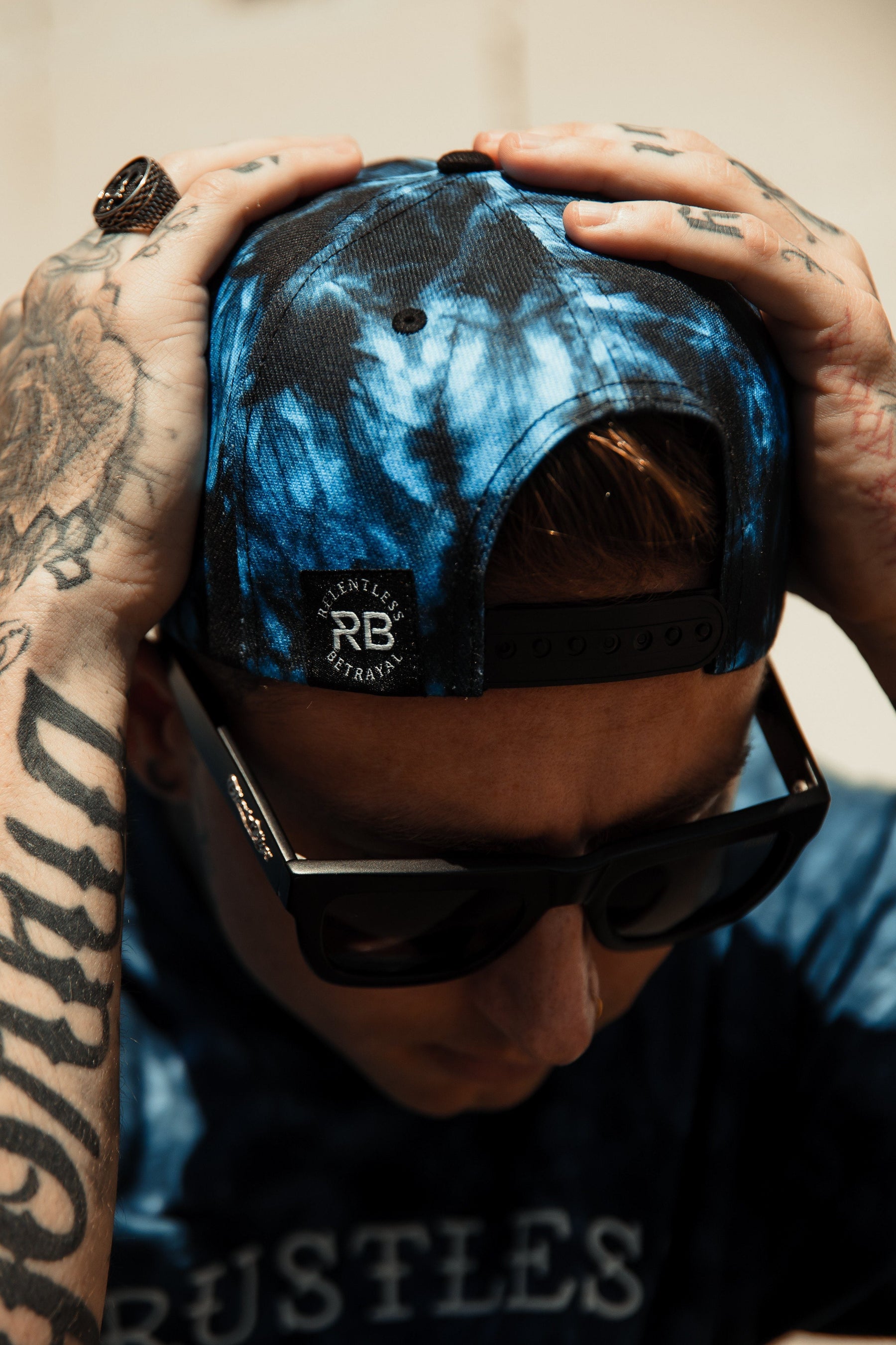 A person in a Lost At Sea tie-dye snapback and sunglasses cradles their tattooed hands over their head. Their shirt features the word RUSTLES, partly visible, while the cap shows a Relentless Betrayal logo, blending unique style and attitude.