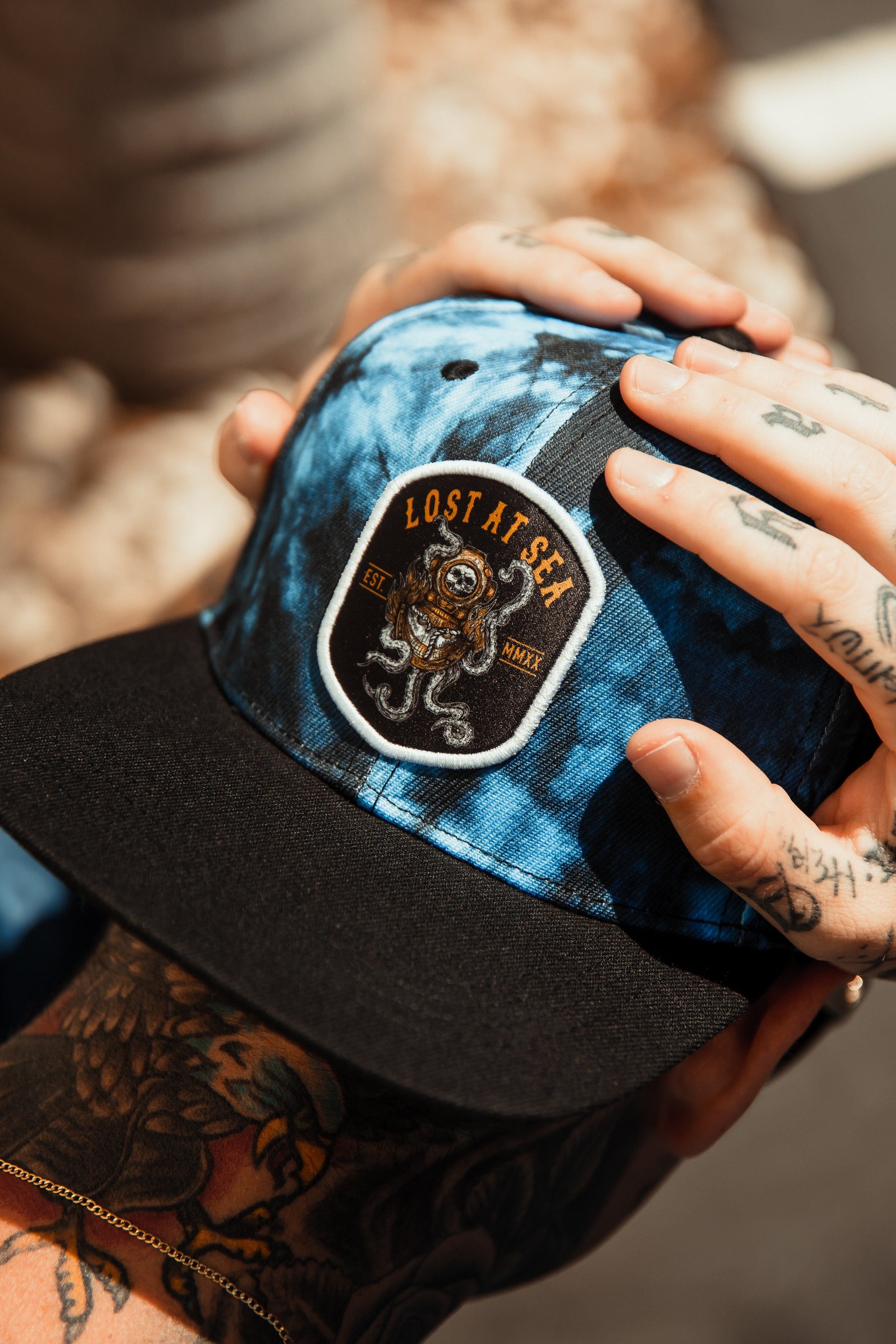 A person with tattooed hands adjusts a Lost At Sea Snapback - Tie Dye by Relentless Betrayal. The hat, featuring a blue-tie dye design with a black brim, showcases an octopus patch and the words LOST AT SEA and EST. MMXL against a blurred background.