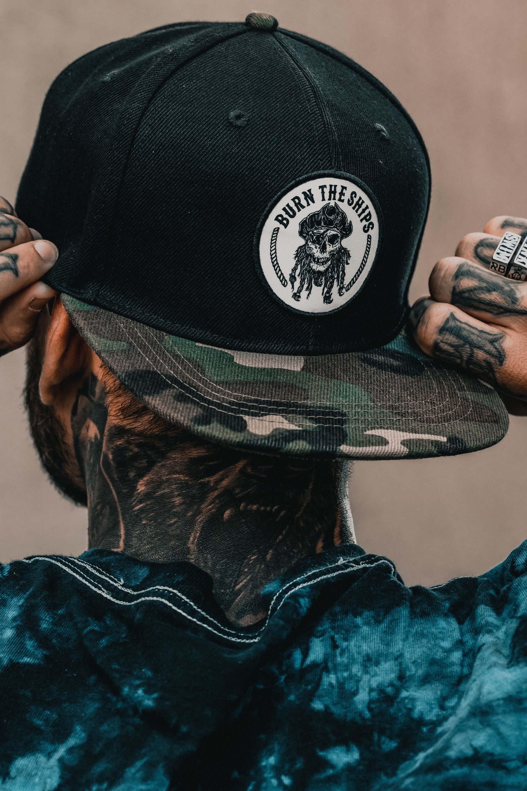 Sporting neck and hand tattoos, a person adjusts the Burn The Ships Camo Snapback by Relentless Betrayal, featuring a Native American chief patch. Their blue tie-dye shirt reflects an adventurous, piratical spirit.
