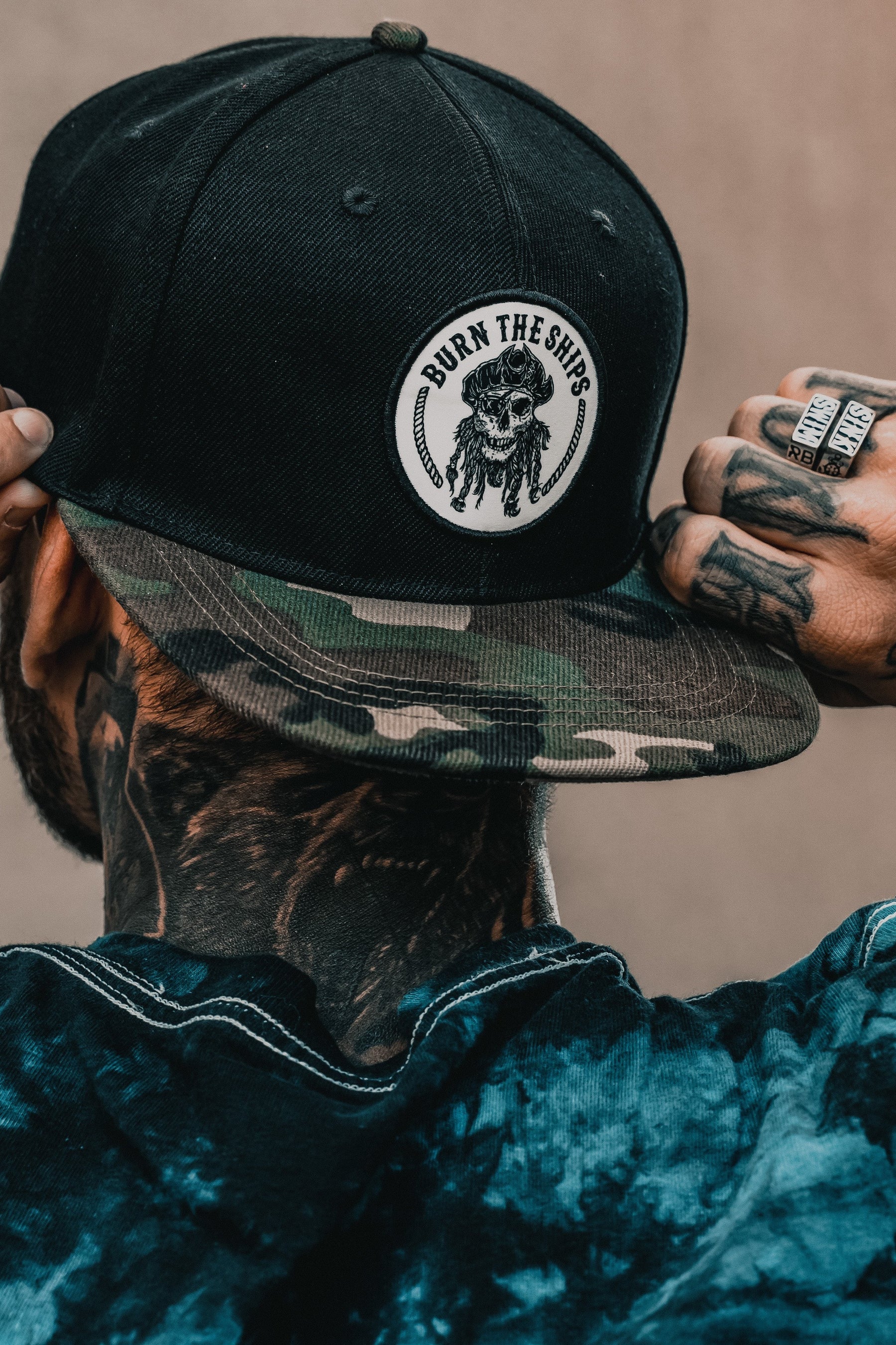 A person adjusts their camo-brimmed cap with the Burn the Ships logo, tattooed hands catching the light. A Relentless Betrayal Sink or Swim Ring glints on one finger, complementing their visible neck tattoos and dark tie-dye shirt.