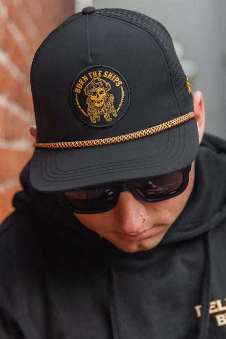 Donning a Relentless Betrayal Burn The Ships BLACKOUT Snapback—a black cap with gold embroidery and a lion logo—they wear sunglasses and a black hoodie against a blurred brick wall, exuding an effortlessly cool vibe.