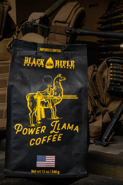The Black Rifle Coffee Power Llama Roast - Ground, with its yellow llama and machine gun design and American flag, sits on rugged terrain with tactical gear. This light roast from Peru Arabica features nutty aromas.