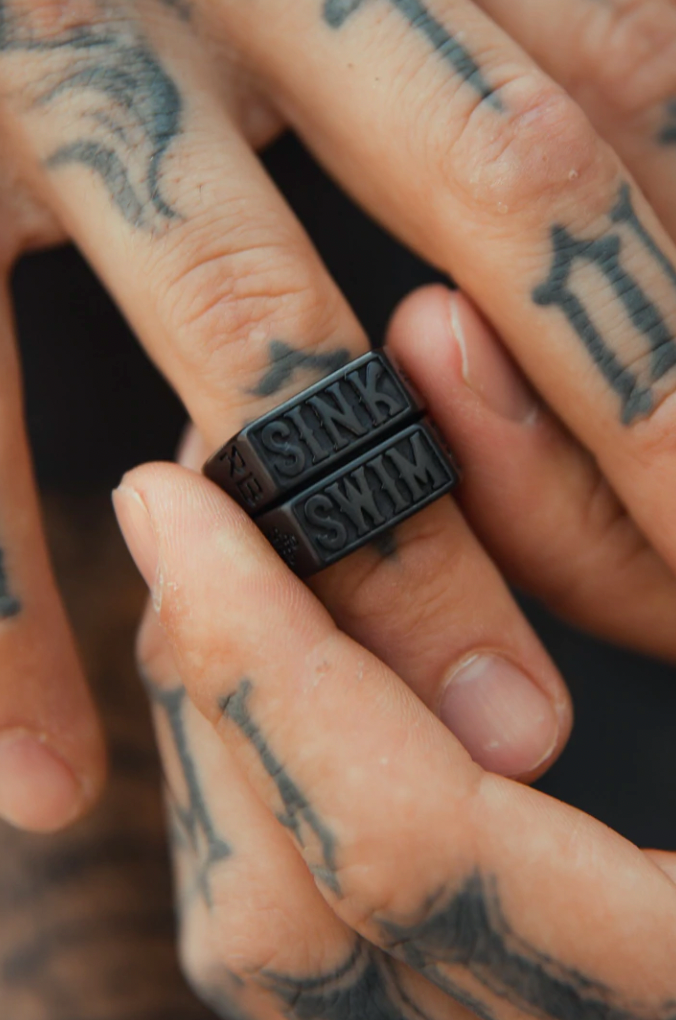 Close-up of tattooed hands showcasing Relentless Betrayals Matte Black Sink or Swim Rings. One ring says SINK while the other, a handcrafted piece, reads SWIM. The tattoos enhance the unique design of this swim jewelry accessory.