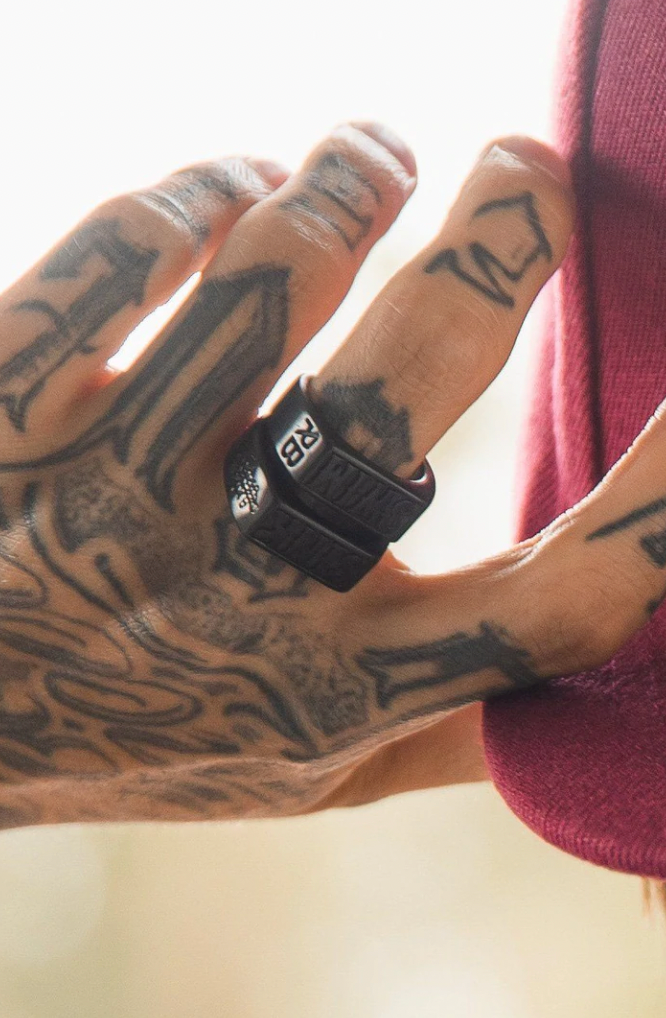 A tattooed hand, adorned with two Matte Black Sink or Swim Rings from Relentless Betrayal, grips pink fabric. Intricate tattoos cover the fingers and hand, while the inscribed stainless steel ring adds an edgy touch to this unique swim jewelry ensemble.