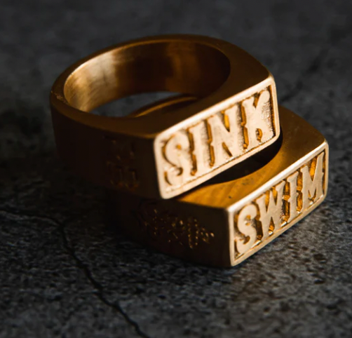 Two handcrafted rings from the Relentless Betrayals Gold Sink or Swim Rings collection rest on a textured surface. One reads SINK and the other SWIM, with their rectangular design and metallic sheen strikingly contrasting against the dark background, highlighting their unique allure.