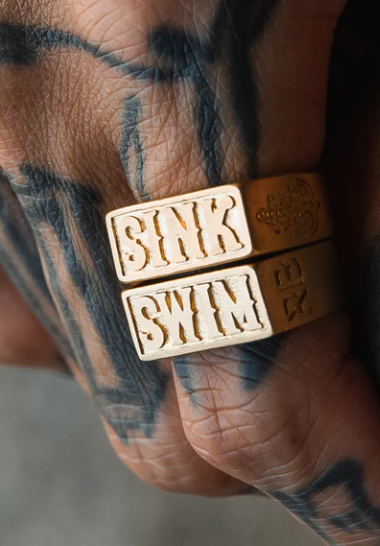 A tattooed hand showcases two Relentless Betrayal Gold Sink or Swim Rings; one engraved with SINK, the other with SWIM. These elegant pieces, bordered by bold tattoos on fingers and knuckles, embody handcrafted artistry reflecting the delicate balance between choice and fate.