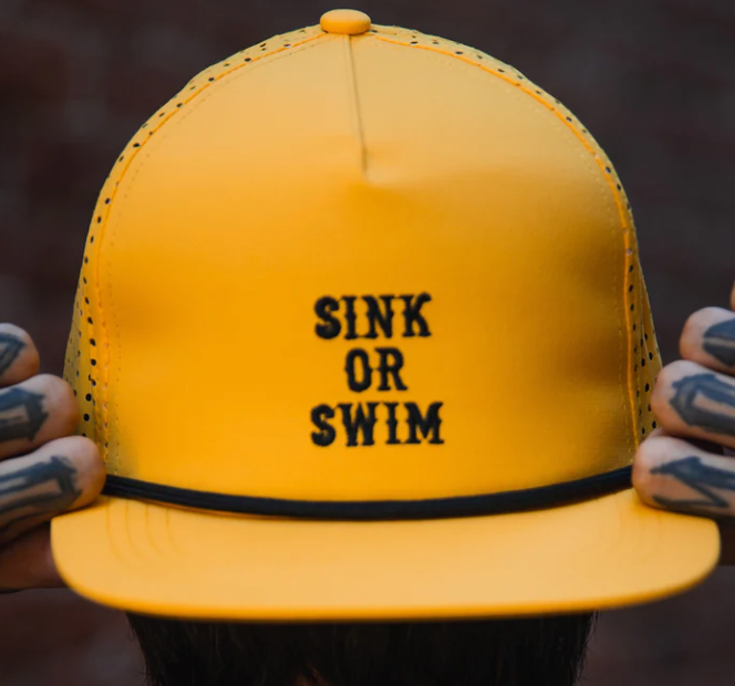 A person with tattooed fingers holds the brim of a bright yellow Gold Sink or Swim Snapback by Relentless Betrayal, featuring SINK OR SWIM in black embroidery. The background is blurred.