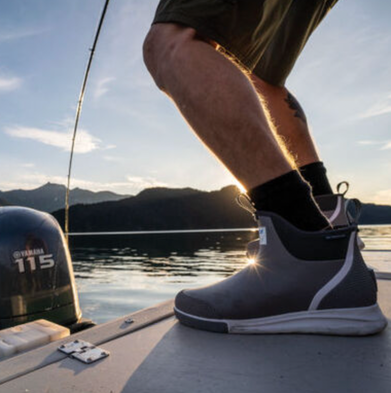 A close-up of a persons leg shows the Xtratuf Mens Ankle Deck Sport boots on a boat deck, featuring a slip-resistant outsole. The scene captures the sun setting over a calm lake with mountains in the distance and a Yamaha motor resting quietly, ideal for an evening of sportfishing.
