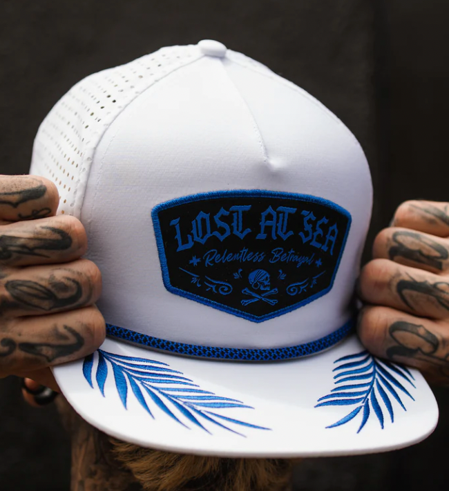 A person with tattooed hands holds the Lost at Sea Whiteout Snapback by Relentless Betrayal. The hat features a blue and black Lost At Sea: Relentless Betrayal patch and a brim printed with blue palm leaves, enhancing its nautical charm.