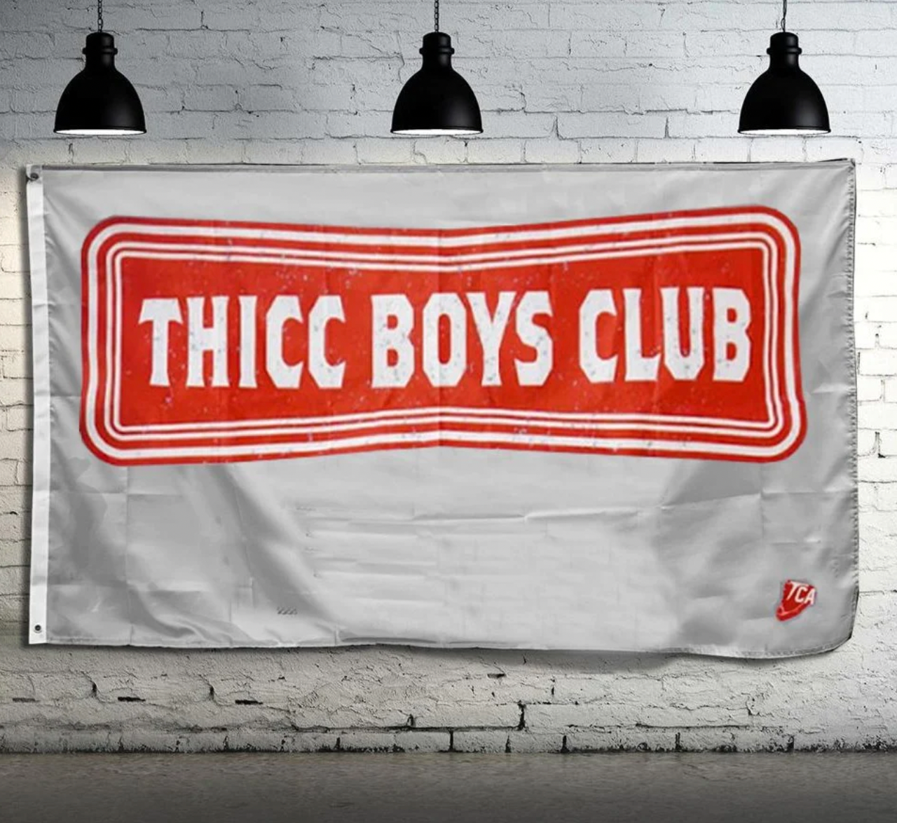 The Team Thicc Boys Club Flag by Tango Charlie Apparel, with a red rectangular THICC BOYS CLUB emblem on white polyester, hangs on a white brick wall. Black industrial lights highlight its double-stitching and metal grommets, while a TCA logo appears in the bottom right corner.