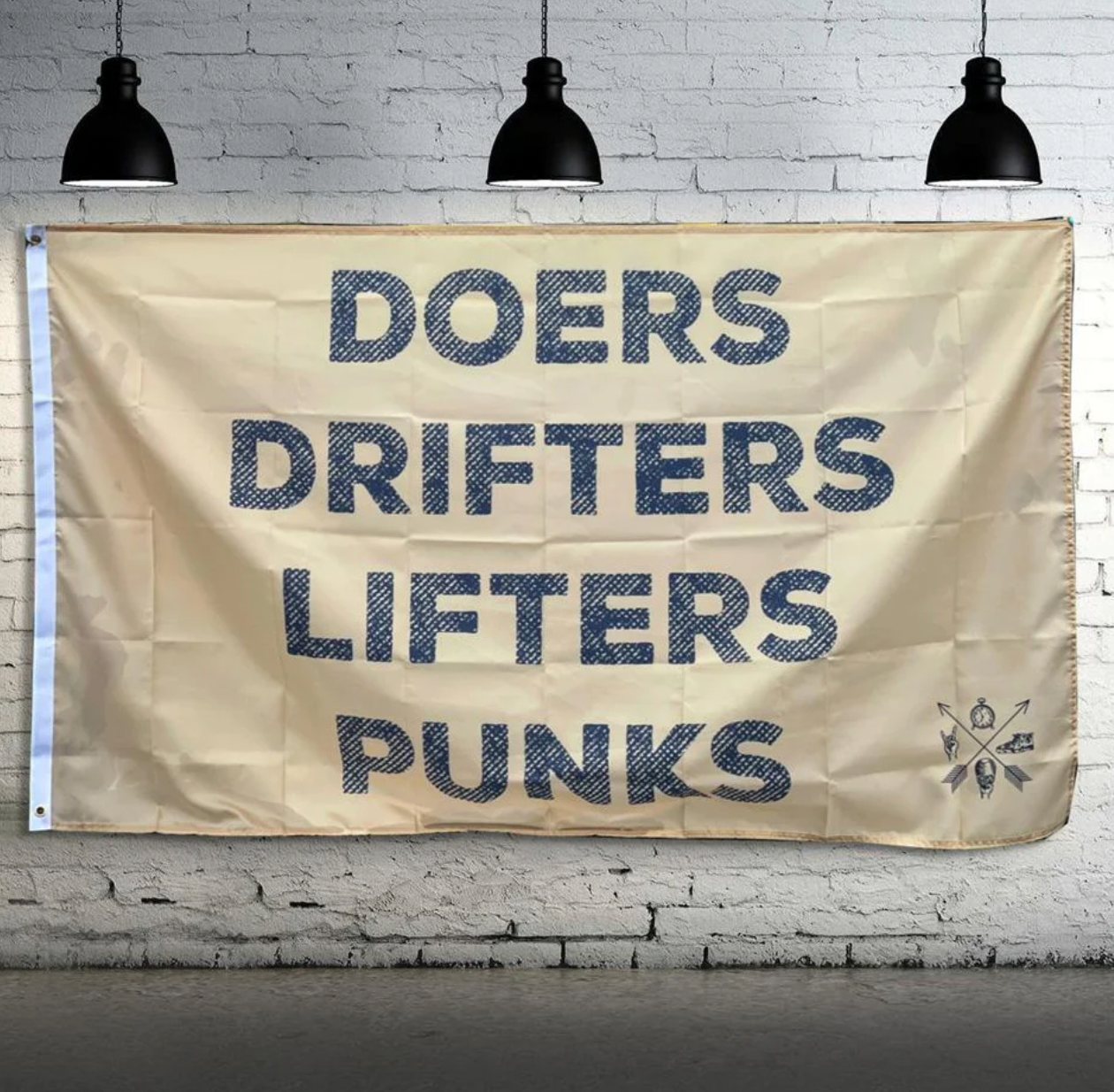 The Team Doers Drifters Lifter Punk Flag by Tango Charlie Apparel features bold text and hangs on a white brick wall. It includes metal grommets and double-stitching, illuminated by three black industrial-style lights above.
