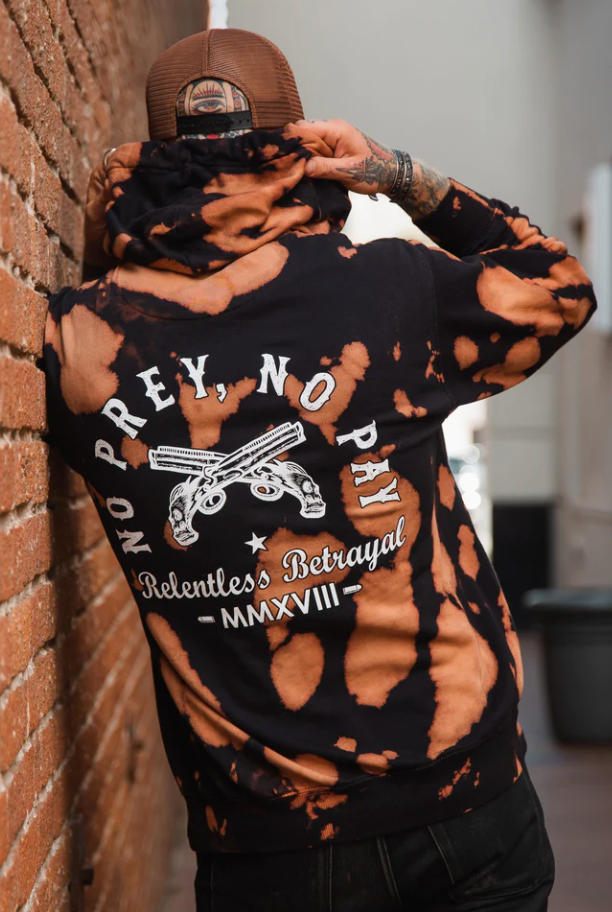 A person in a backward cap leans against a brick wall, wearing the Relentless Betrayal No Prey, No Pay bleach wash hoodie. This unisex hoodie features two crossed guns and the text No Prey, No Pay, Relentless Betrayal, MMXVIII on the back, made from a cozy cotton polyester blend fleece.