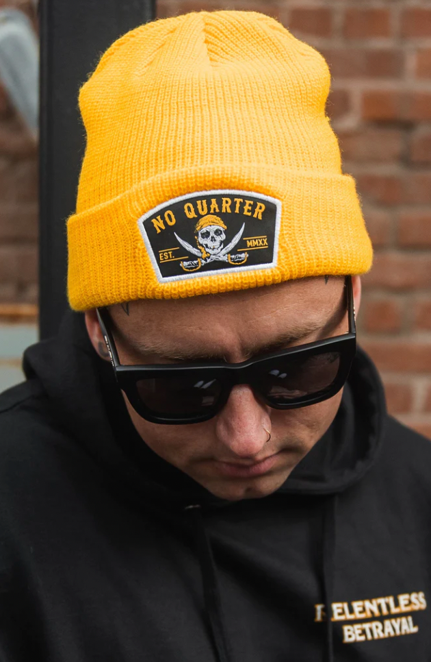 Wearing a No Quarter vibrant yellow beanie with a skull and crossbones patch, sunglasses, a nose ring, and a Relentless Betrayal black hoodie, they pose coolly against a rugged brick wall.