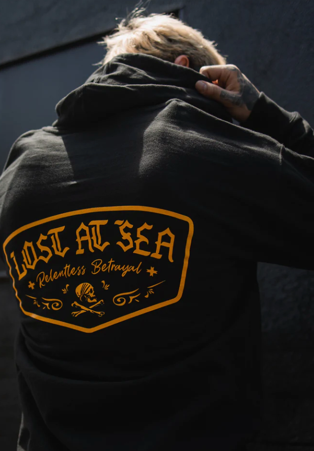 A blond-haired person faces away wearing the Lost at Sea Hoodie by Relentless Betrayal, made from a comfy cotton/polyester blend. It features LOST AT SEA and Relentless Betrayal text with a skull and crossbones graphic in a unisex, edgy, timeless style.